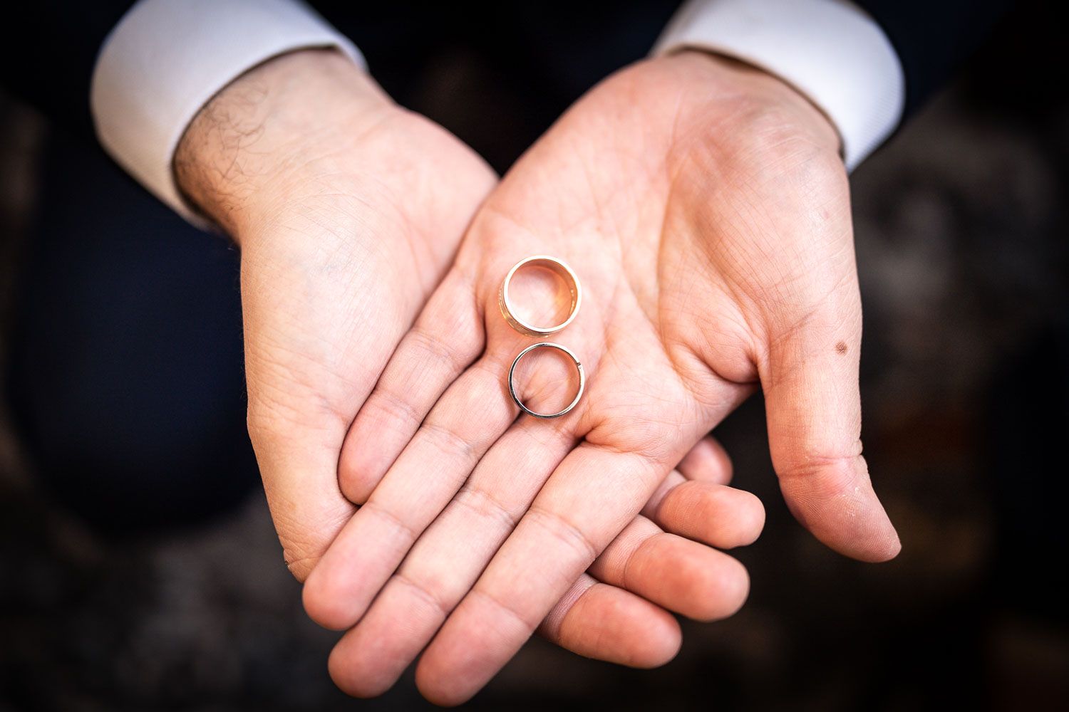 Two open palms holding gold wedding bands against a dark background.