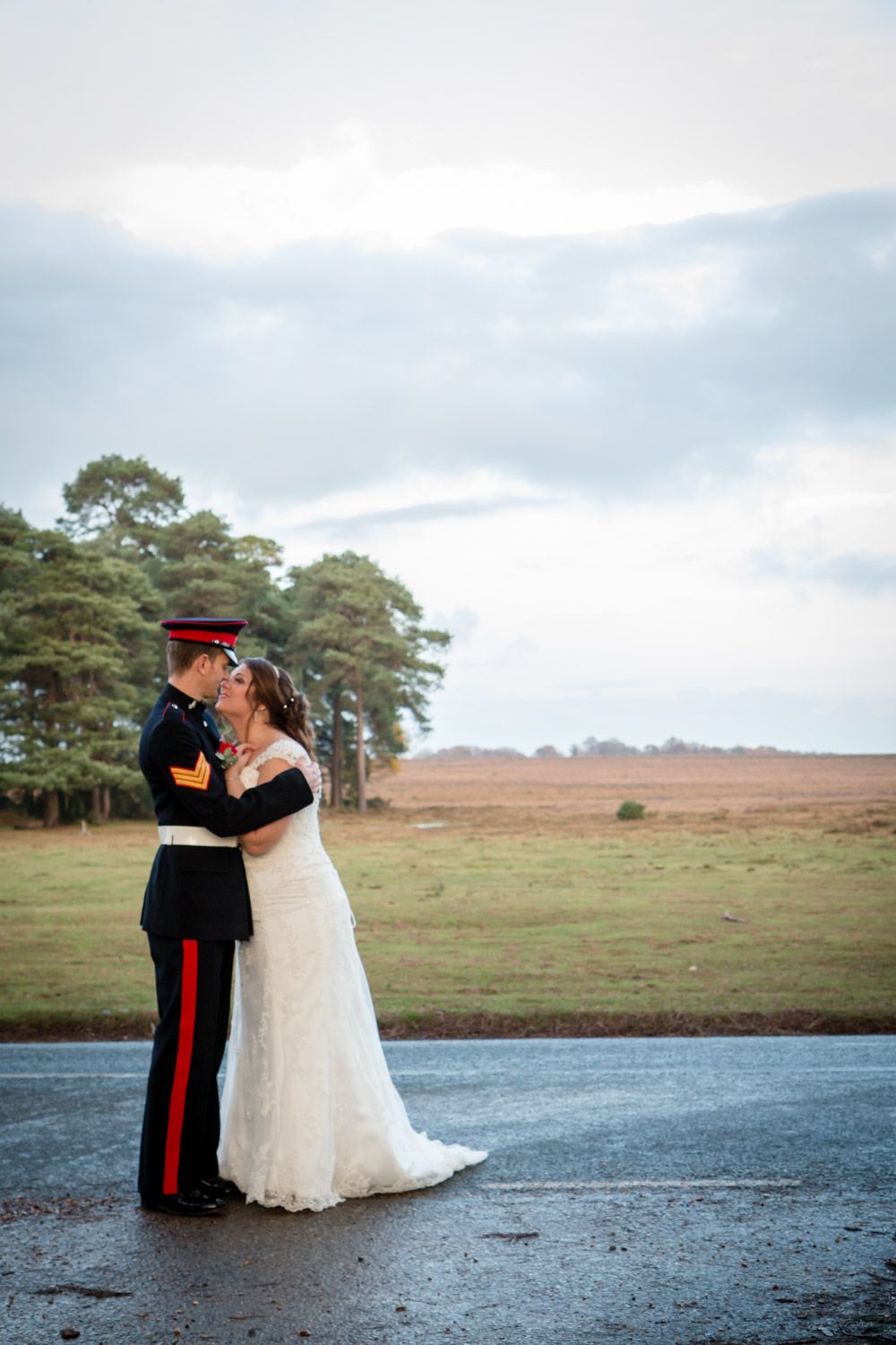 A couple embraces on a wet road surrounded by pine trees and open fields under a cloudy sky.