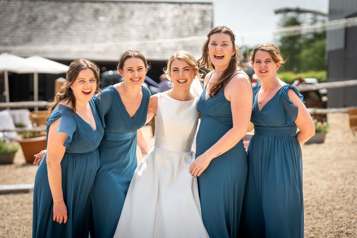 Bride in white gown smiling with four bridesmaids in teal blue dresses at an outdoor wedding venue.