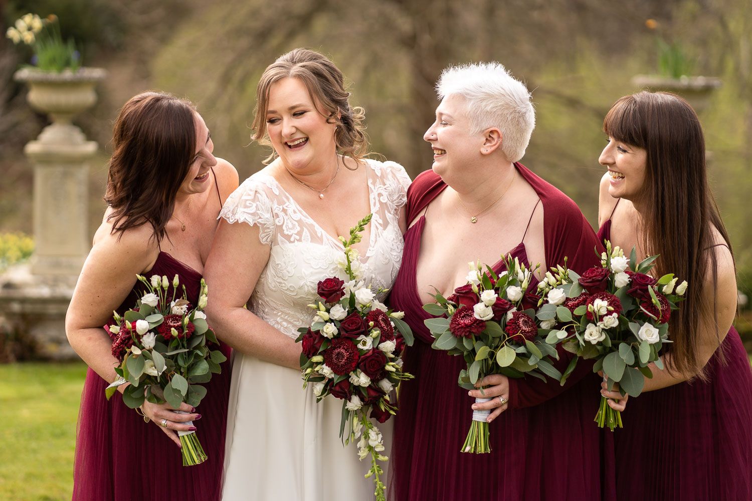 Wedding party holding burgundy and white floral bouquets share joyful laughter in outdoor garden setting.