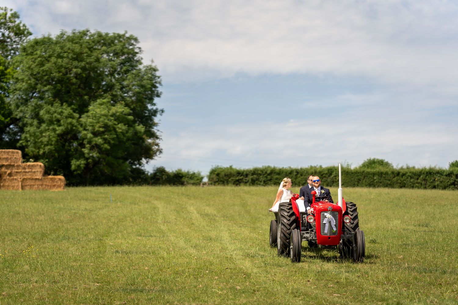Bride and groom riding a red tractor across a green field on their wedding day, with hay bales in the background.