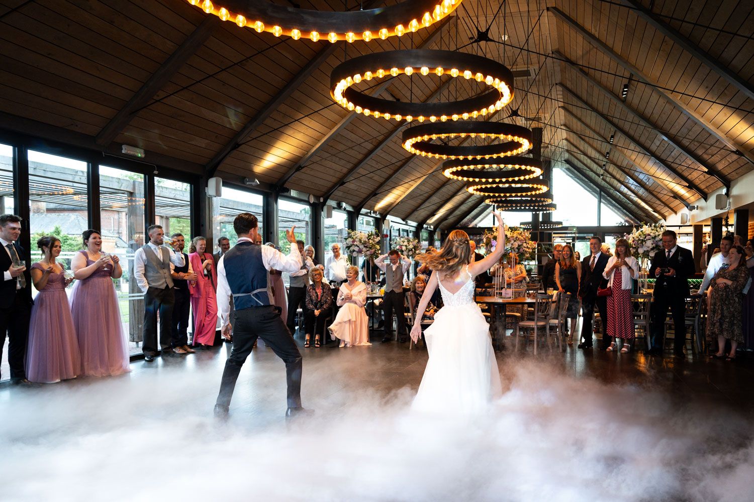A couple shares their first dance surrounded by guests under round string lights with fog effects at a rustic reception venue.