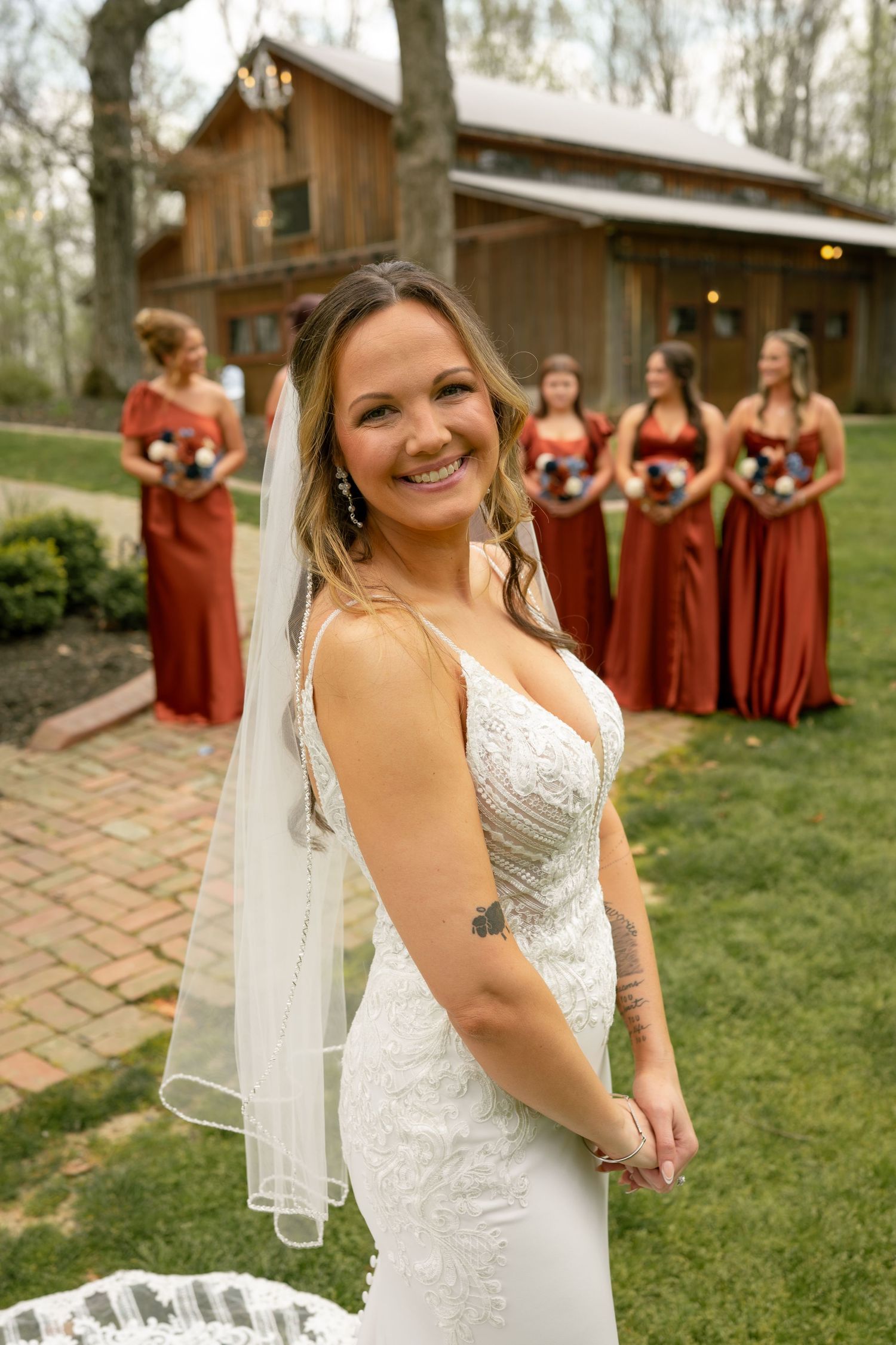 Smiling bride in white lace gown with veil poses outdoors, bridesmaids in rust dresses holding bouquets near rustic barn.