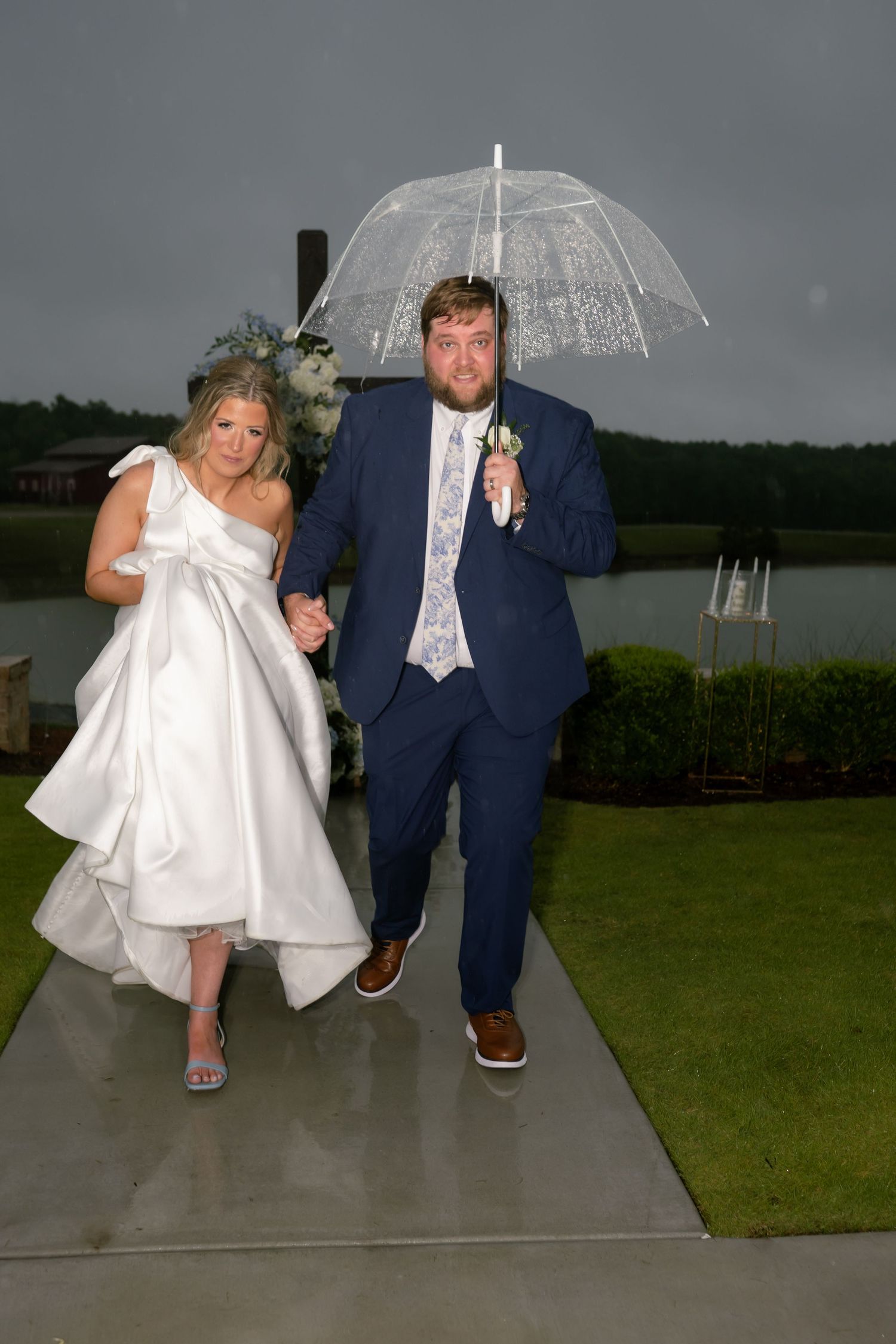 Newlyweds walking under clear umbrella in rain after outdoor lakeside wedding ceremony.