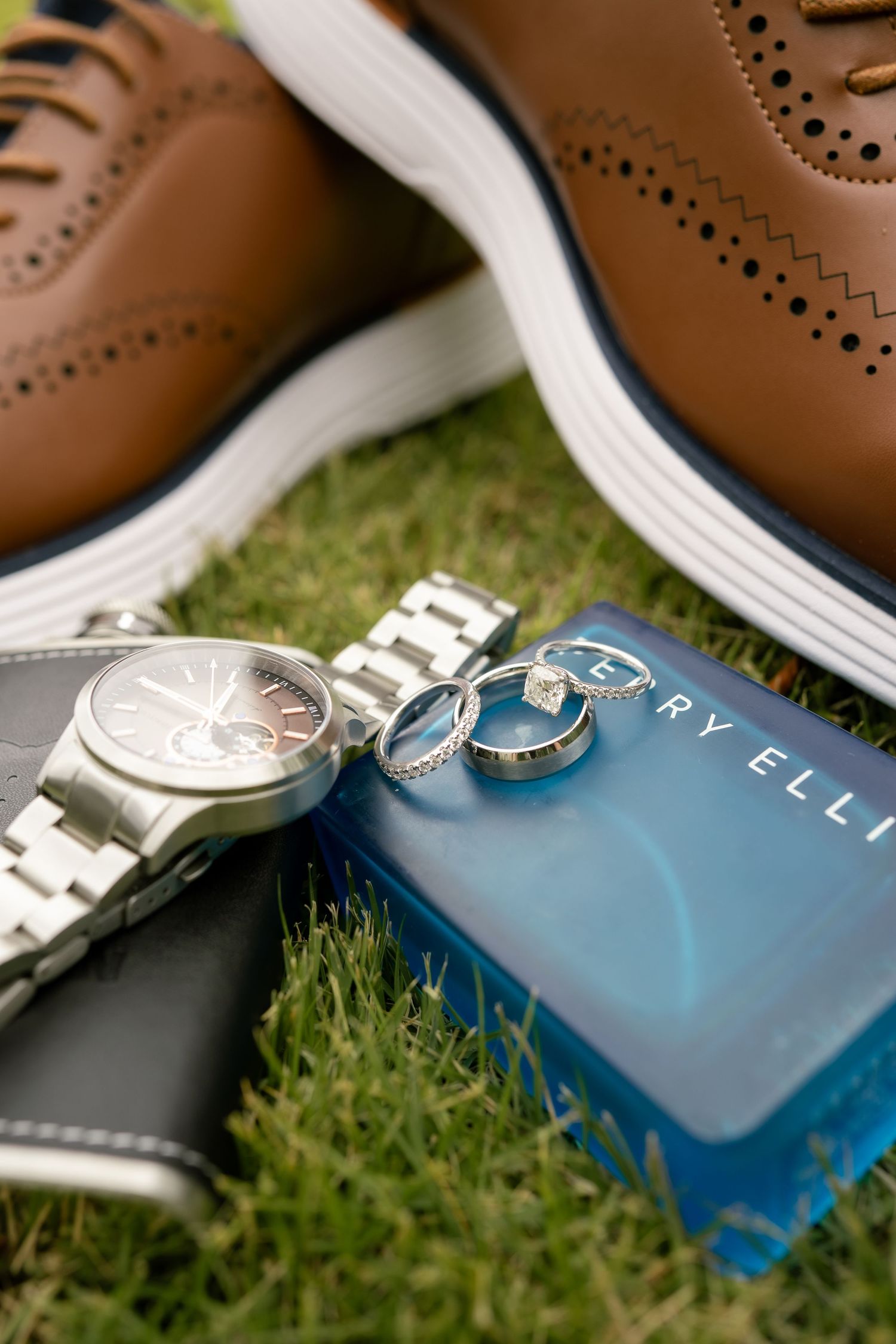 Groom's wedding accessories flat lay featuring brown brogue shoes, silver watch, wedding rings, and Perry Ellis cologne on grass.