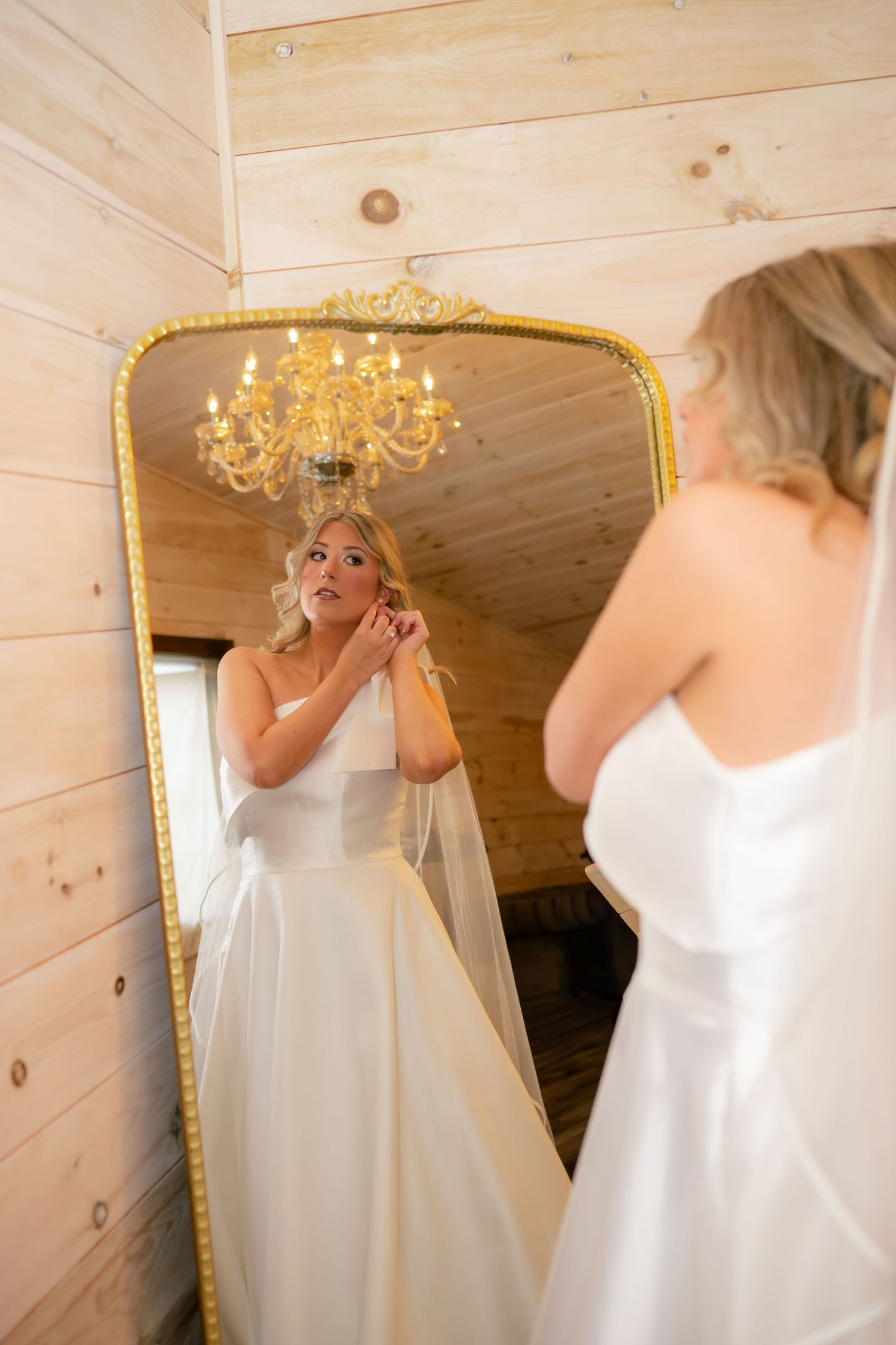 Blonde bride in white gown adjusting earring while looking in ornate gold mirror.