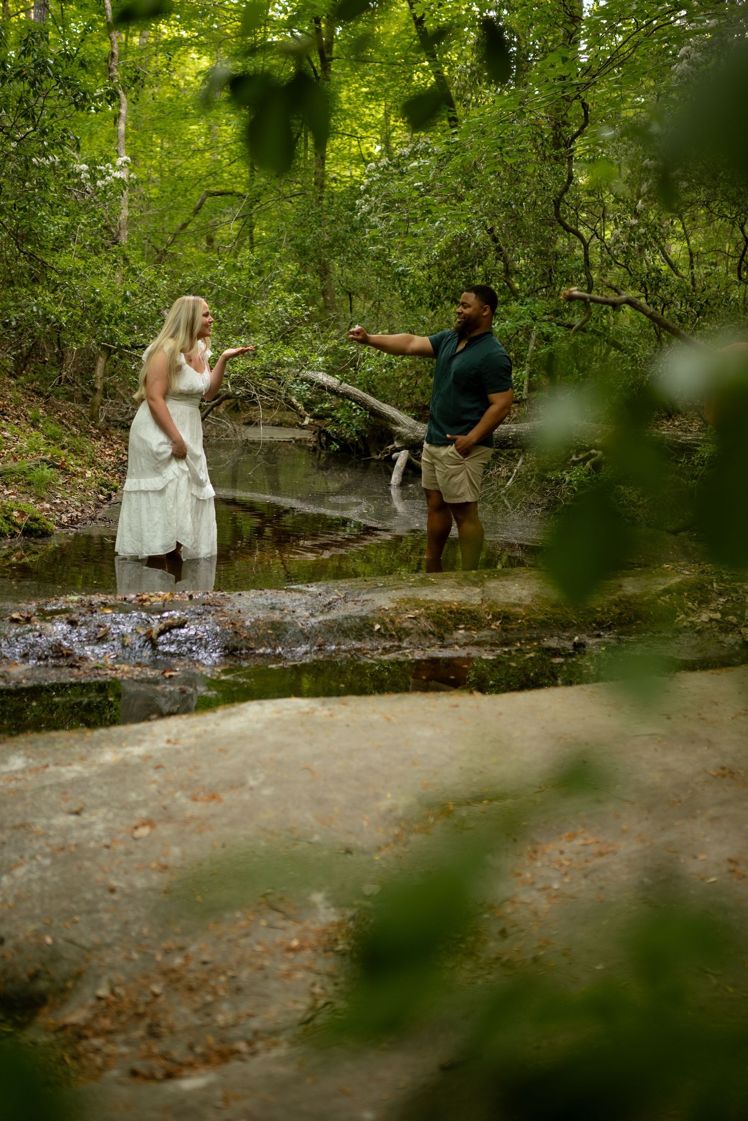 A couple stands in a forest creek, man in green shirt pointing while woman in white dress wades in shallow water.