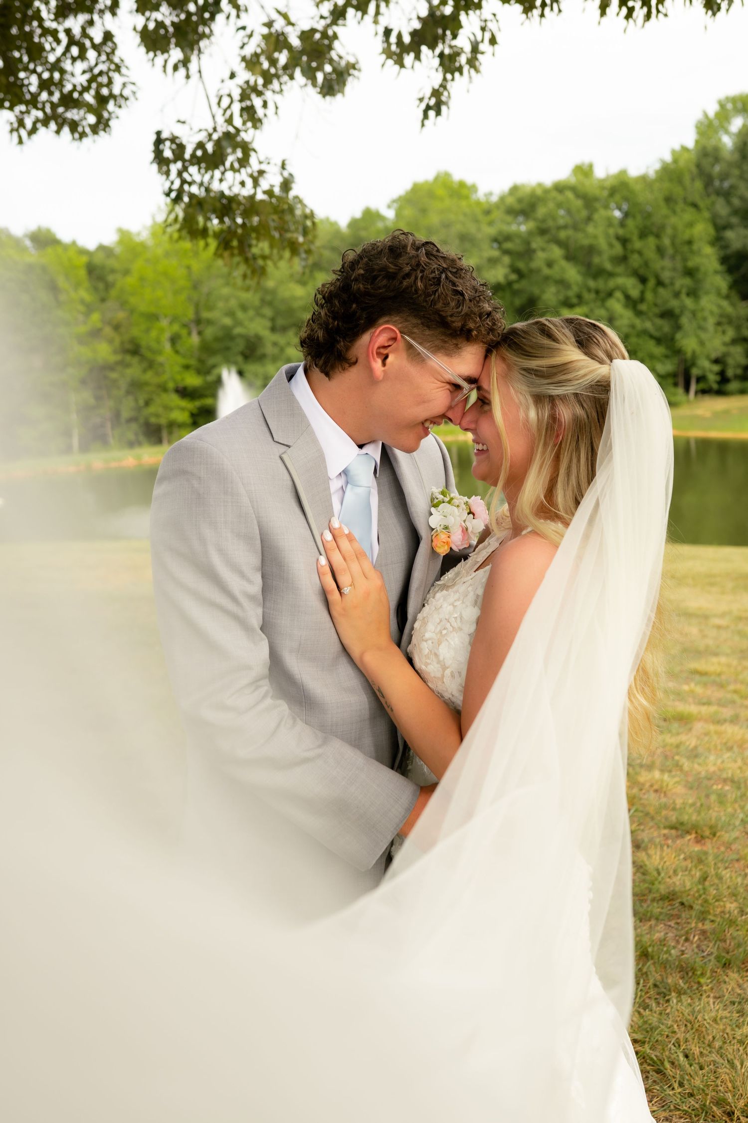 Bride and groom sharing a romantic moment under a tree with a veil flowing around them at an outdoor wedding.