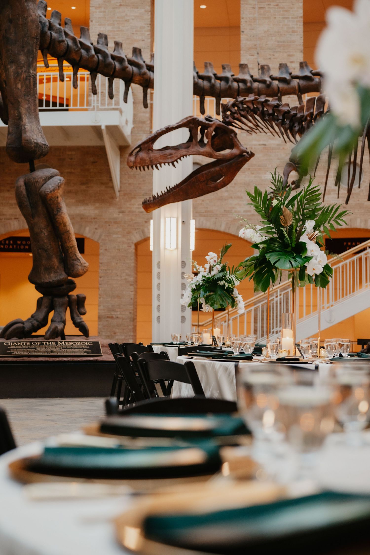 Elegant table setting beneath dinosaur skeleton display in museum atrium with hanging floral arrangements.