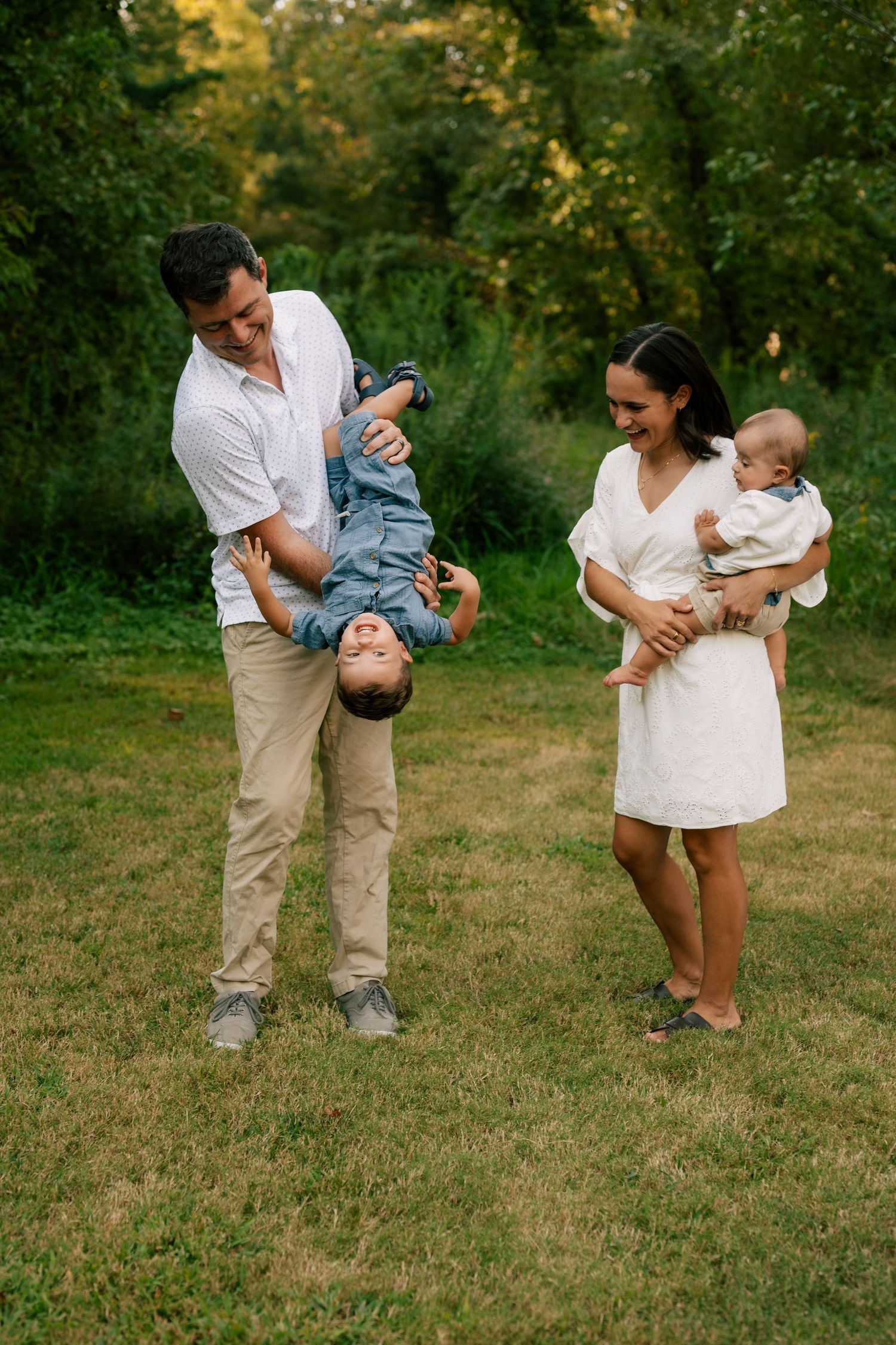 A family plays and laughs together on a grassy lawn during a summer outdoor photoshoot.