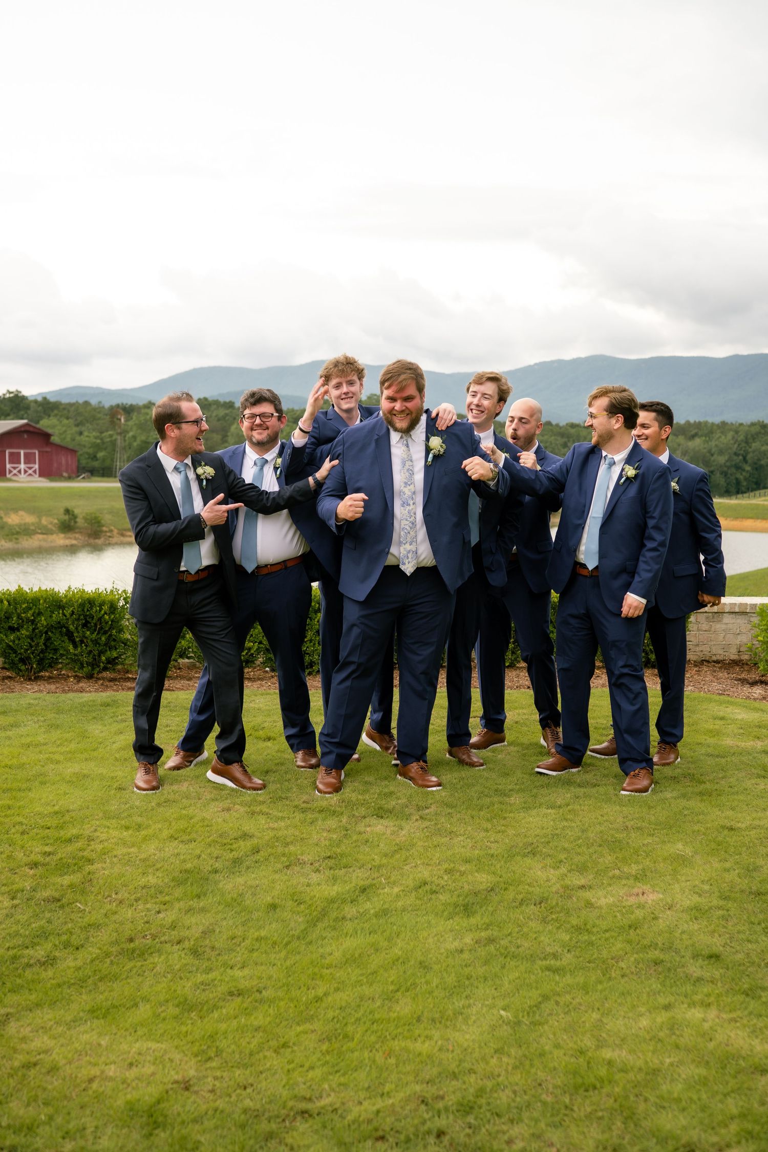 Groom and groomsmen in navy suits laughing and celebrating outdoors with mountain and pond views in background.
