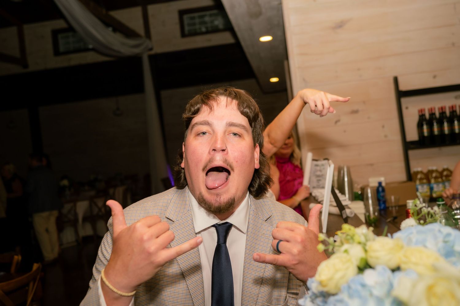 Excited wedding guest in tan suit making fun gestures at reception celebration.