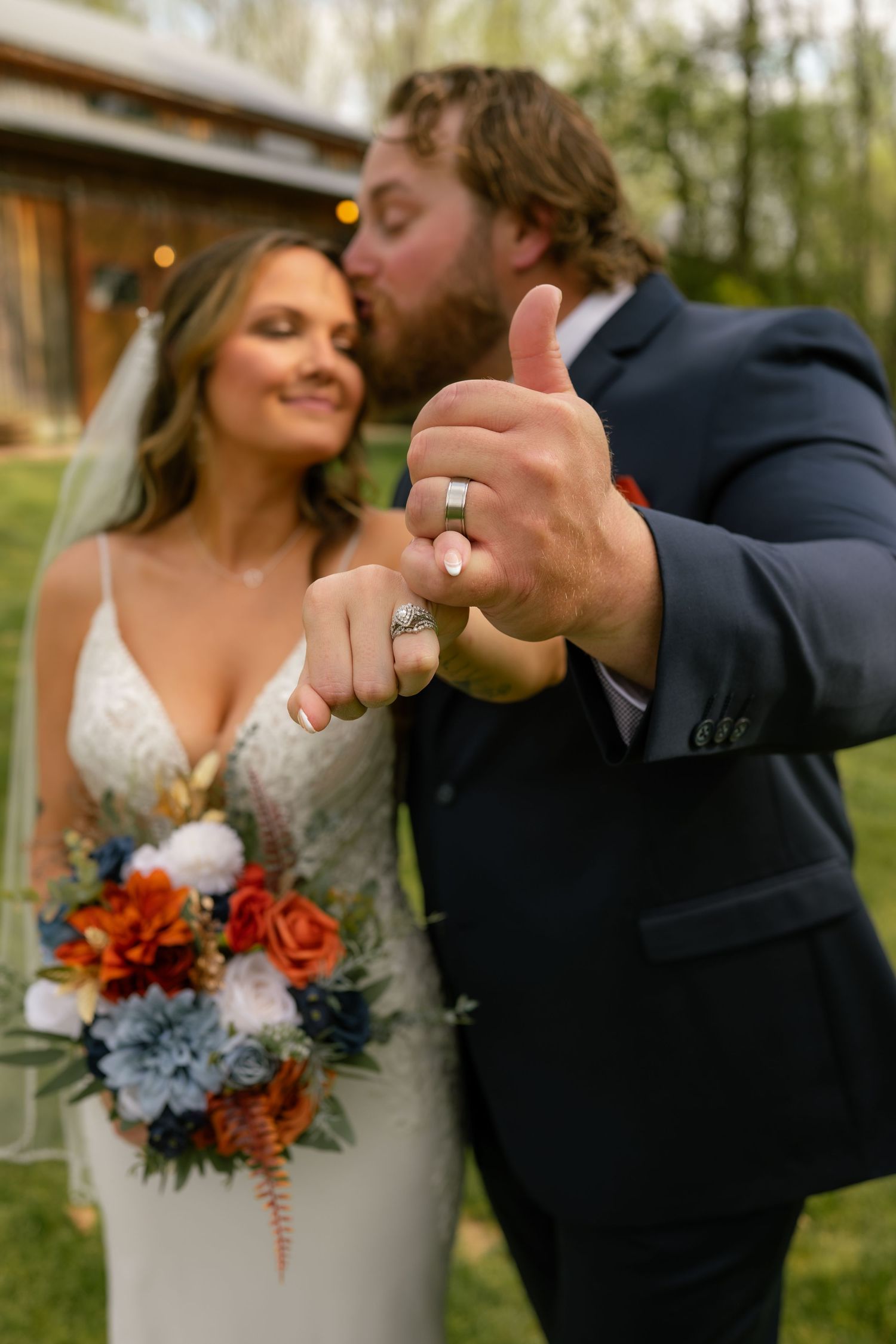 Newlyweds give thumbs up showing wedding rings, bride holds rust and navy bouquet, groom in navy suit.