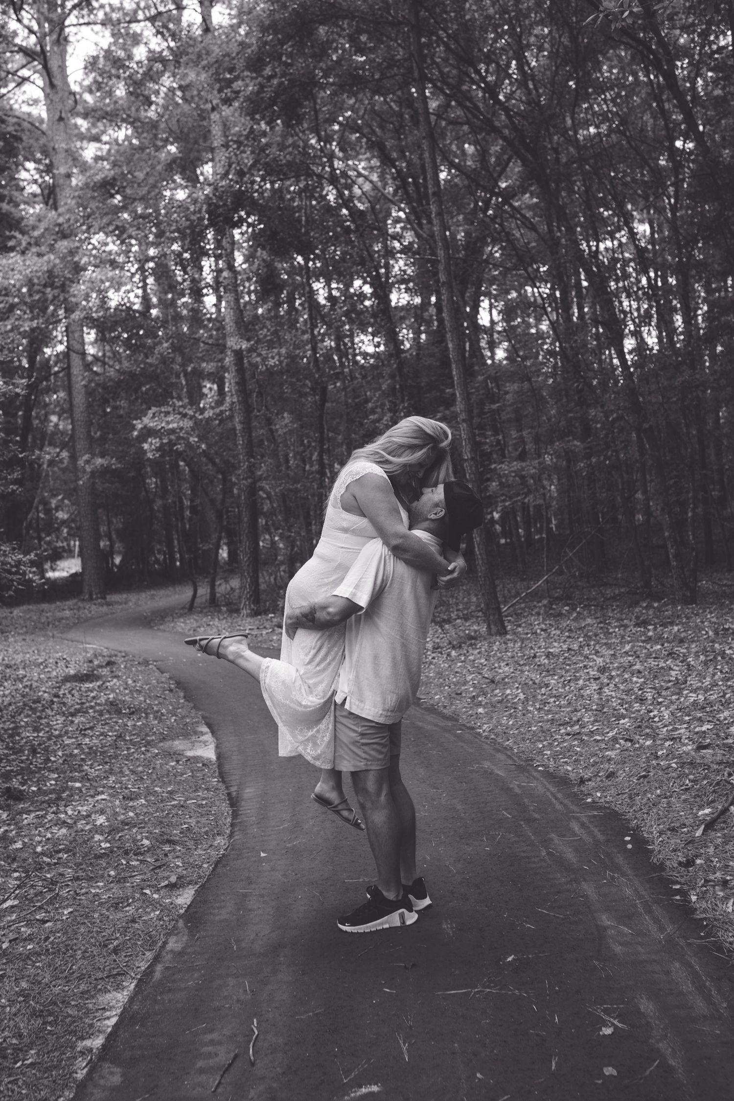Black and white photo of a couple embracing and kissing on a forest path, woman in white dress lifted off ground.