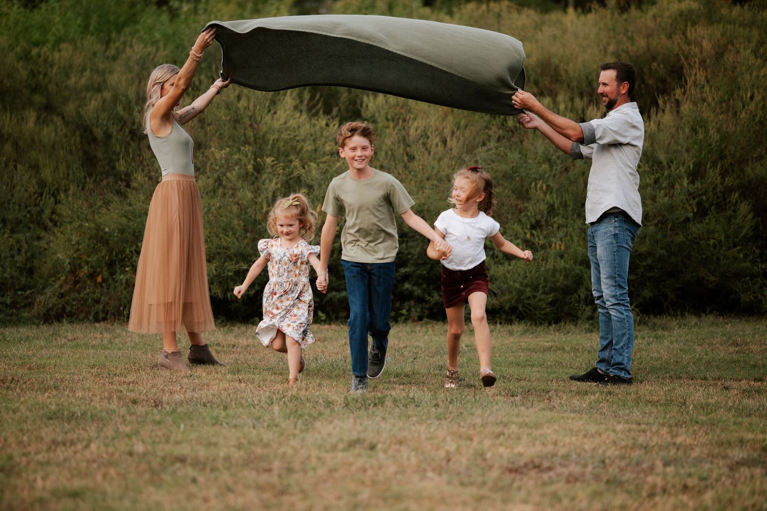 Family plays together outdoors holding a blanket in the air while children run underneath.