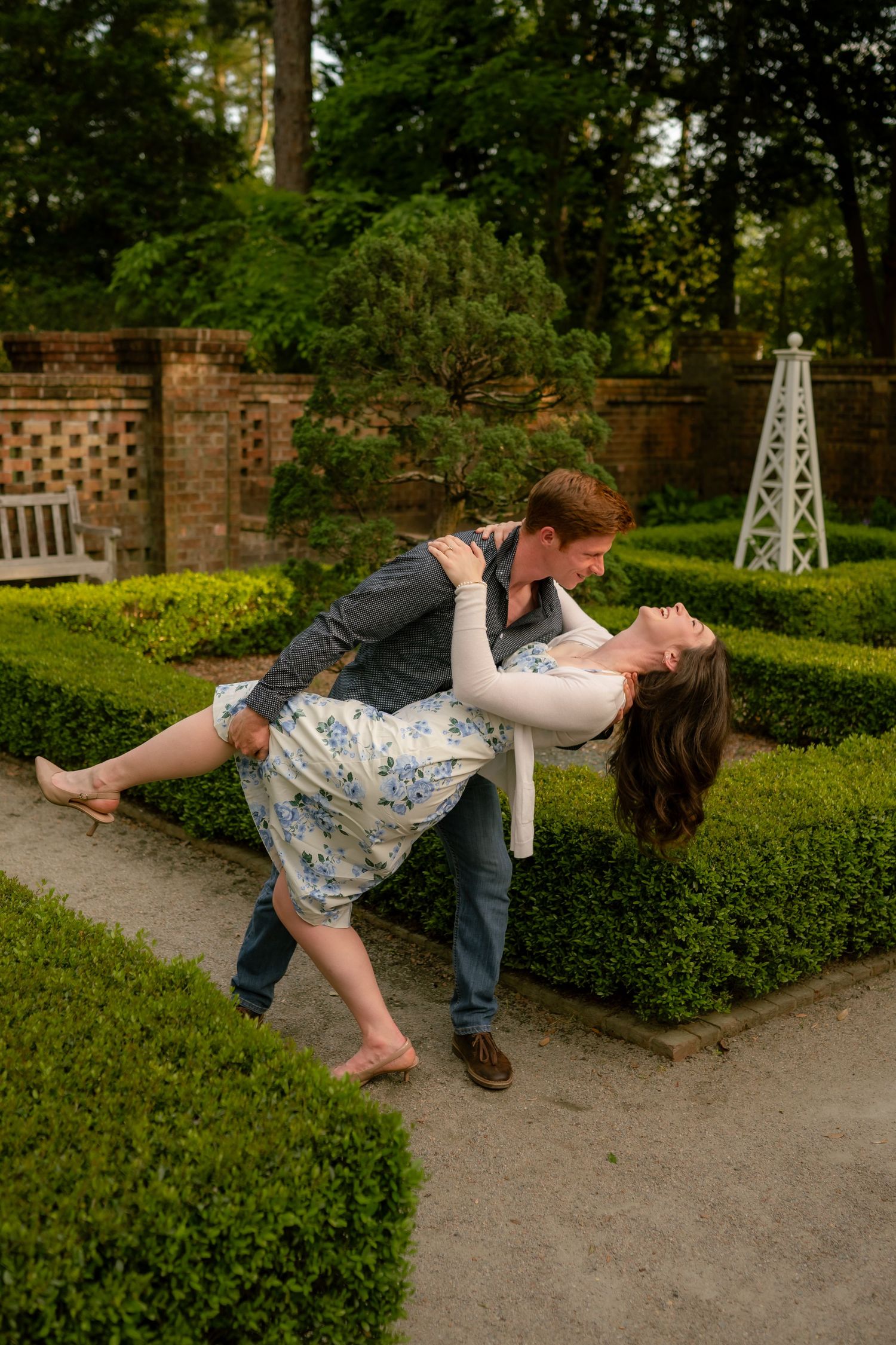 Couple sharing a romantic dip pose in a lush garden with manicured hedges and brick walls.