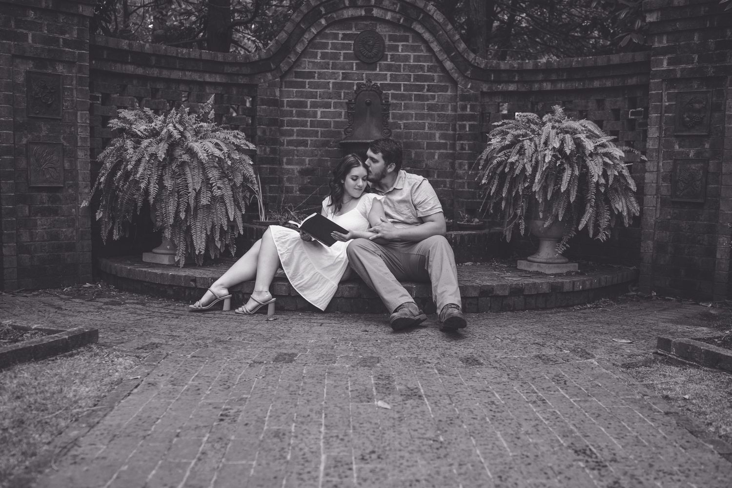 Black and white photo of a romantic couple sitting together reading a book outdoors near a brick wall with ferns.