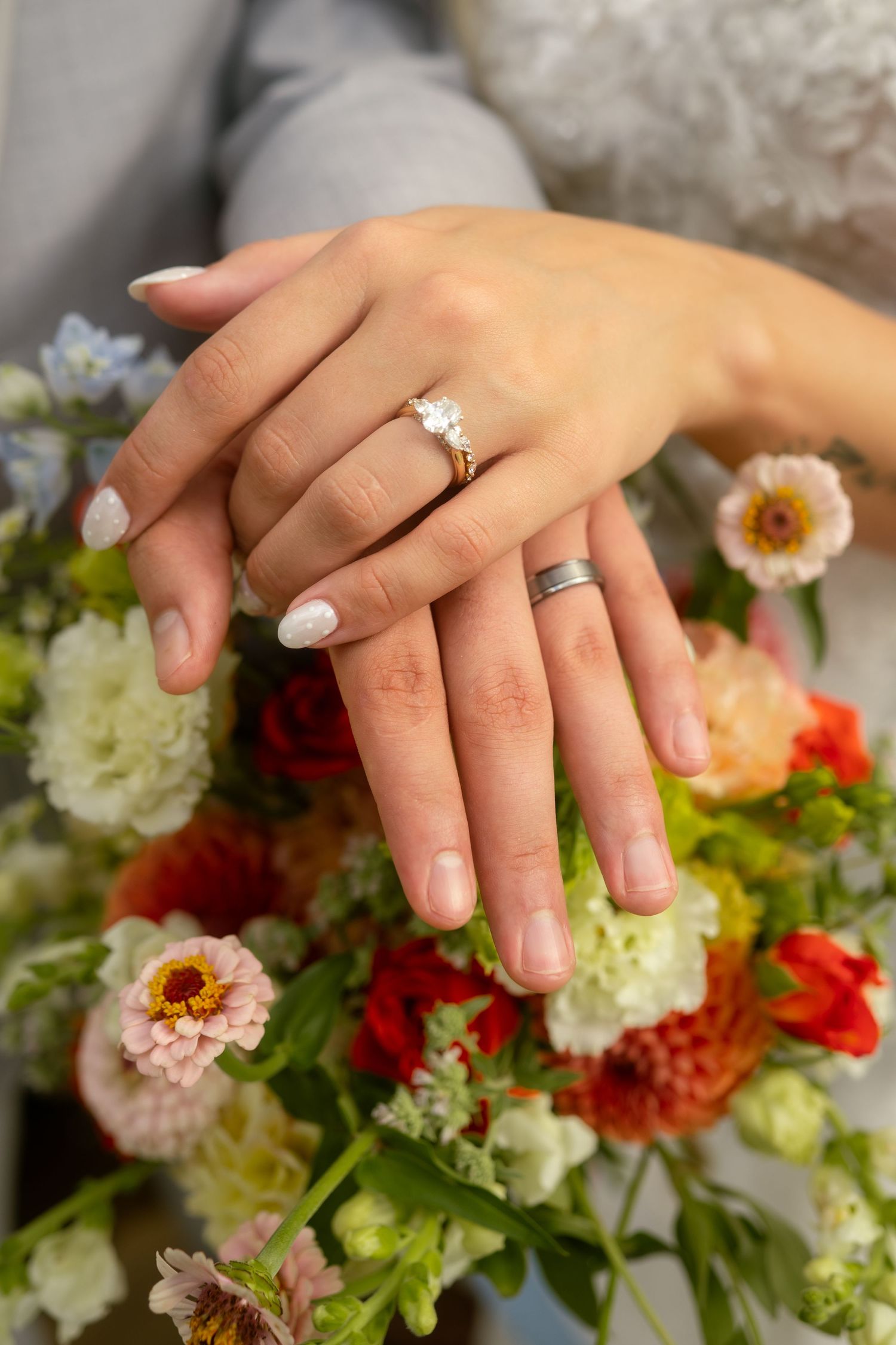 Close-up of bride and groom hands showing diamond engagement ring and wedding band resting on colorful floral bouquet.
