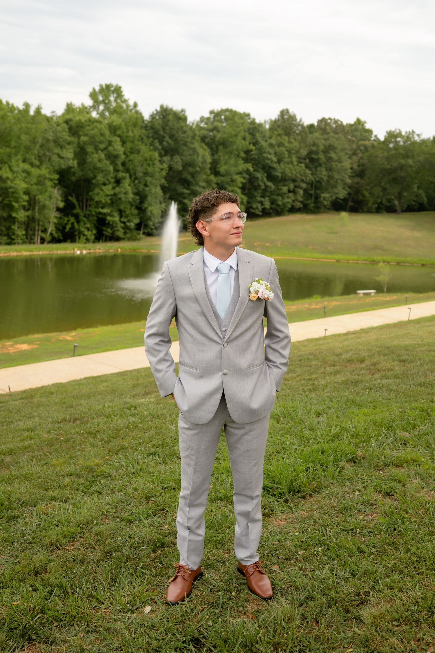 Young groomsman in grey suit with boutonniere stands on green lawn near pond with fountain at wedding venue.