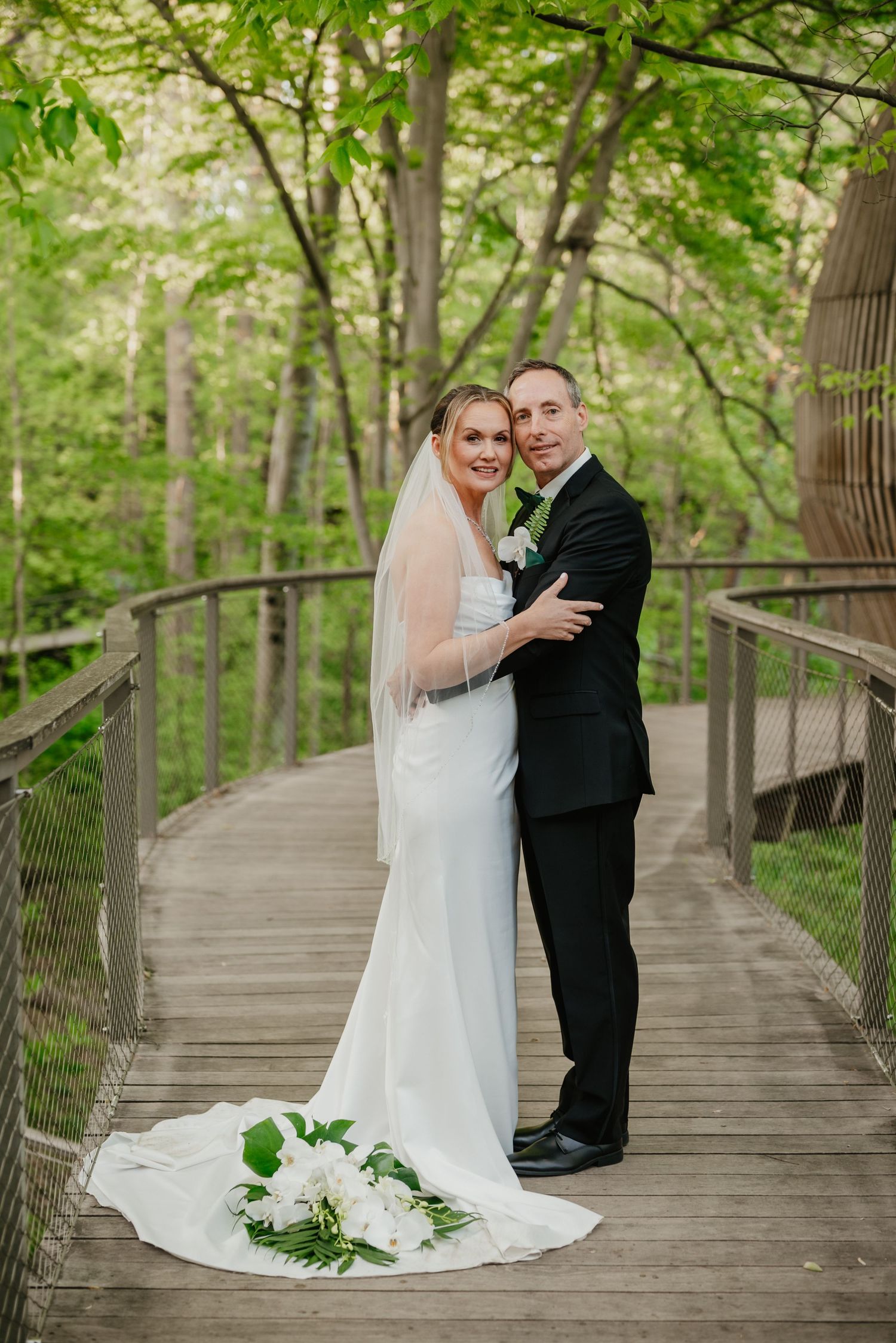 A couple poses on a wooden walkway surrounded by lush green trees on their wedding day.