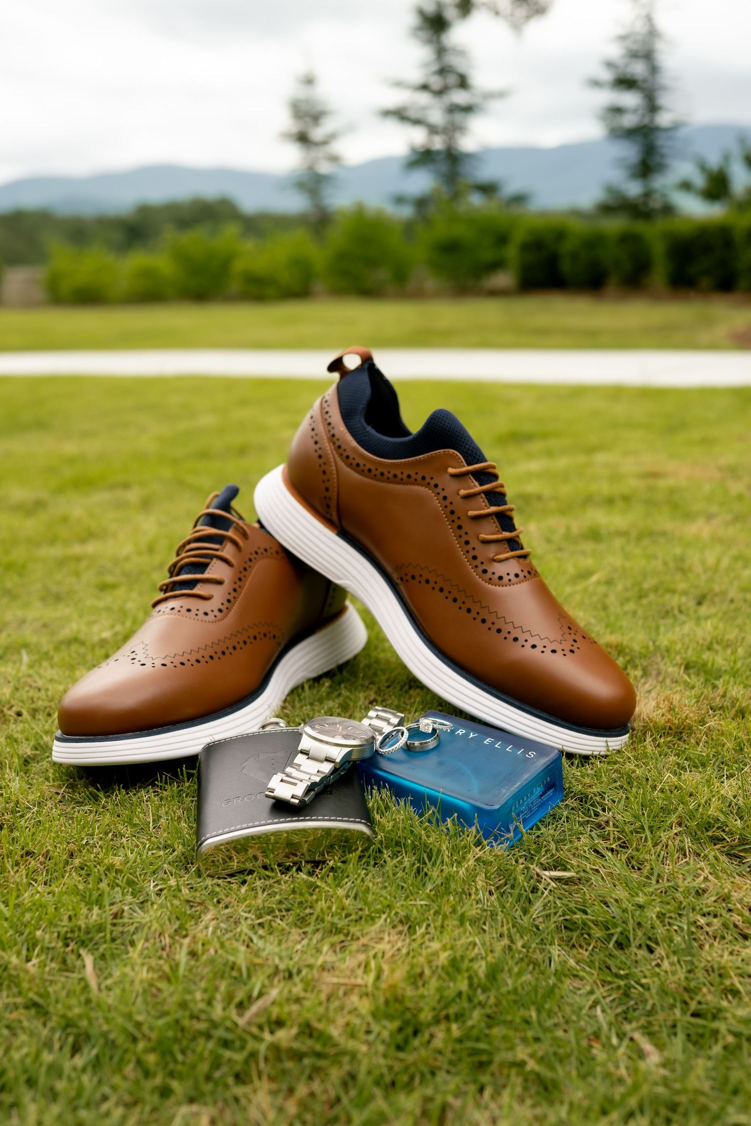 Brown leather Cole Haan wingtip shoes on grass with a watch and accessories nearby.
