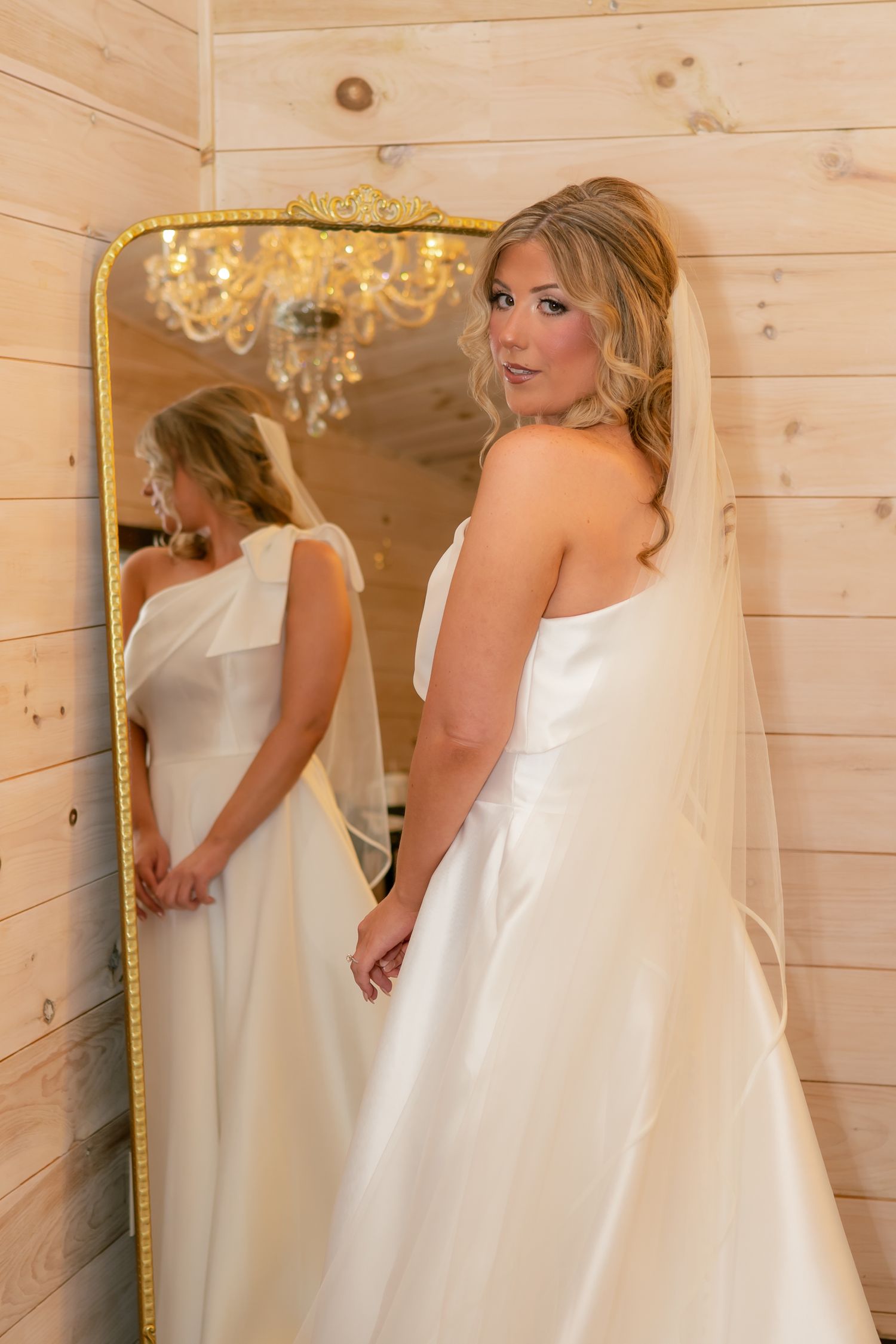 Bride in white gown and veil poses beside gold ornate mirror in wood-paneled room.
