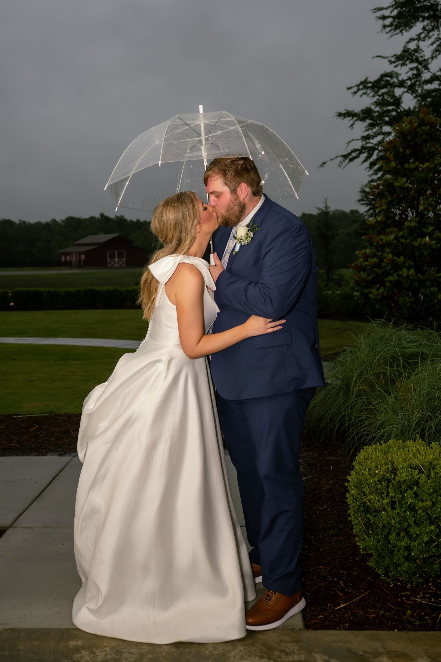 Bride and groom share a romantic kiss under a clear umbrella in the rain outdoors.