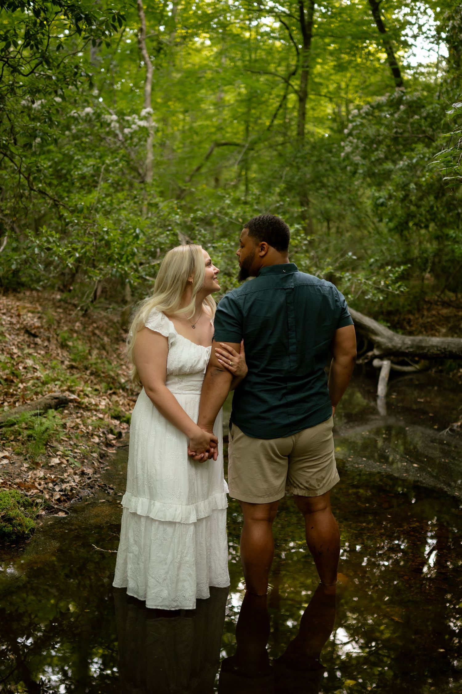 Couple standing in a forest creek, woman in white dress and man in dark shirt holding hands, surrounded by lush green trees.