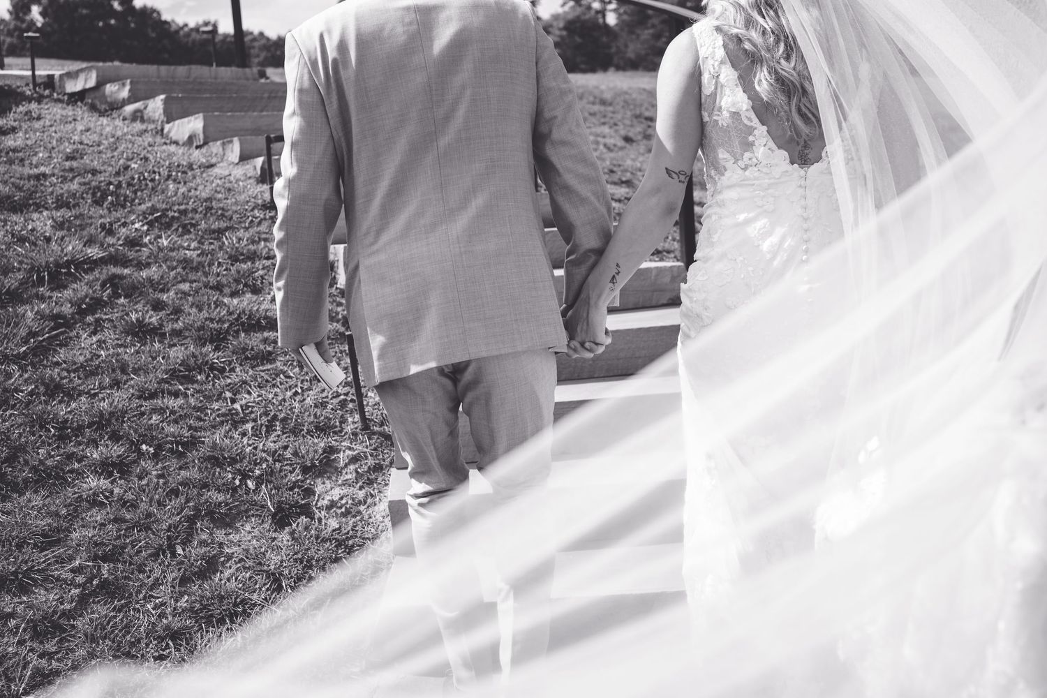 Black and white photo of bride and groom holding hands walking along path with flowing veil at wedding.