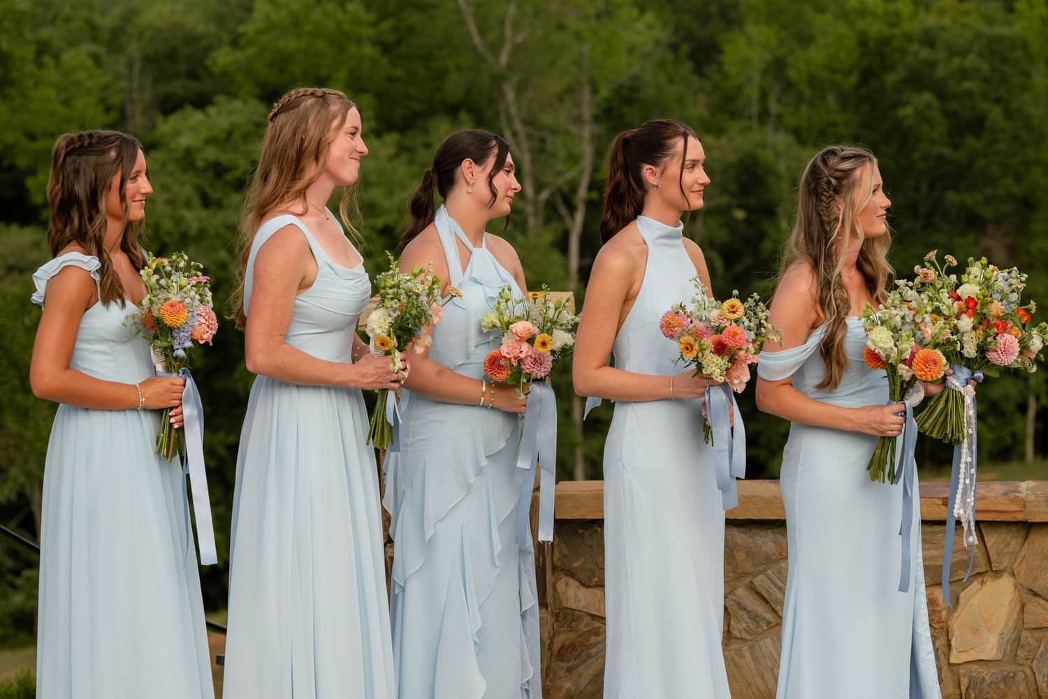 Five bridesmaids in light blue gowns holding orange and white bouquets stand in a row outdoors at a wedding ceremony.