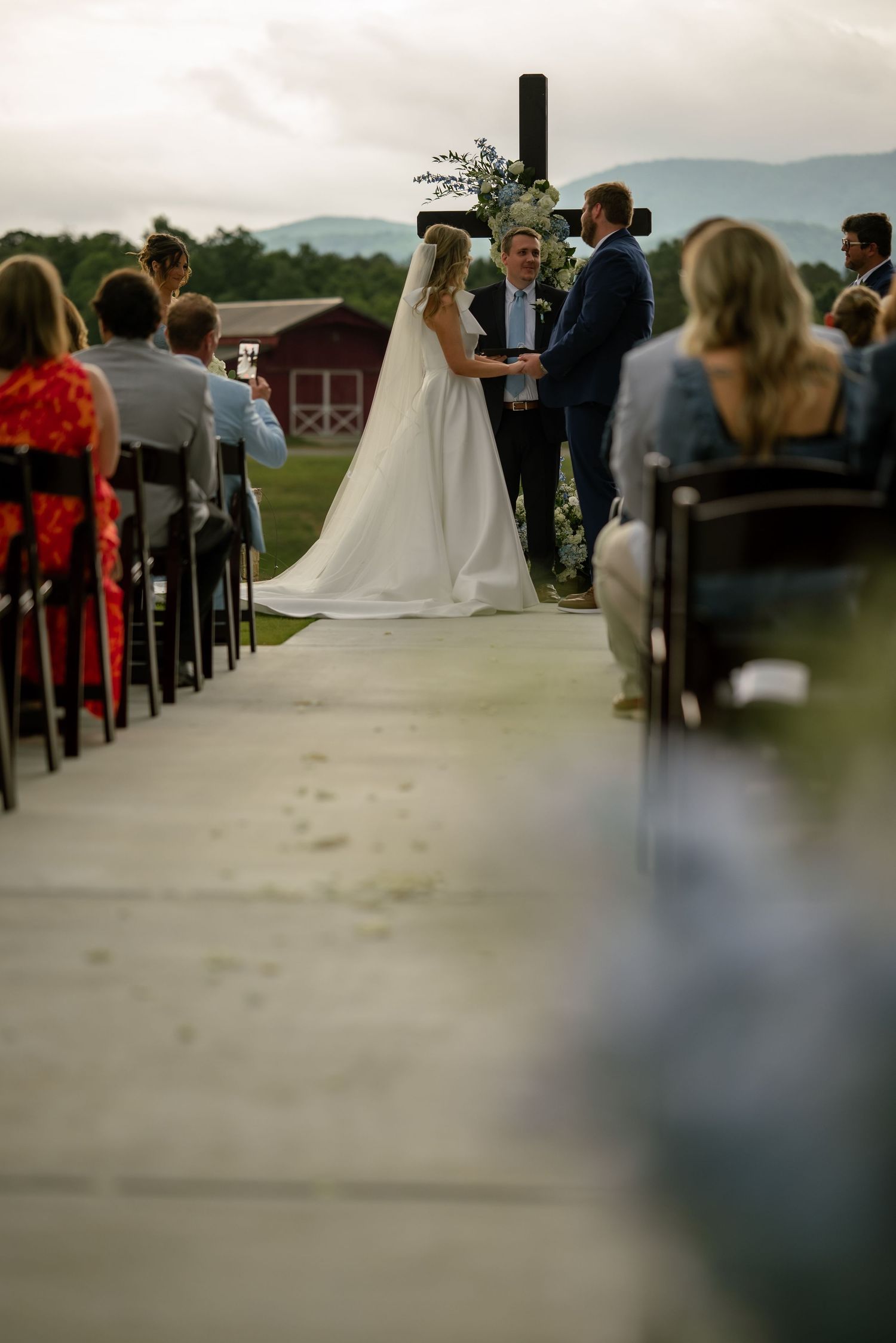 Bride and groom exchanging vows at outdoor ceremony before a wooden cross with mountain views and seated guests.