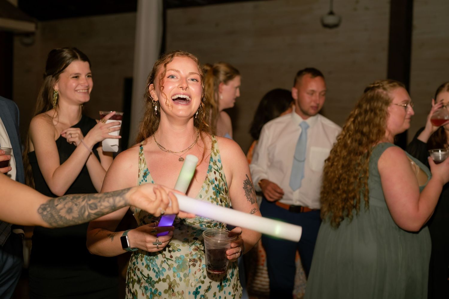 Joyful wedding guest laughs while holding a glow stick during lively outdoor evening reception.