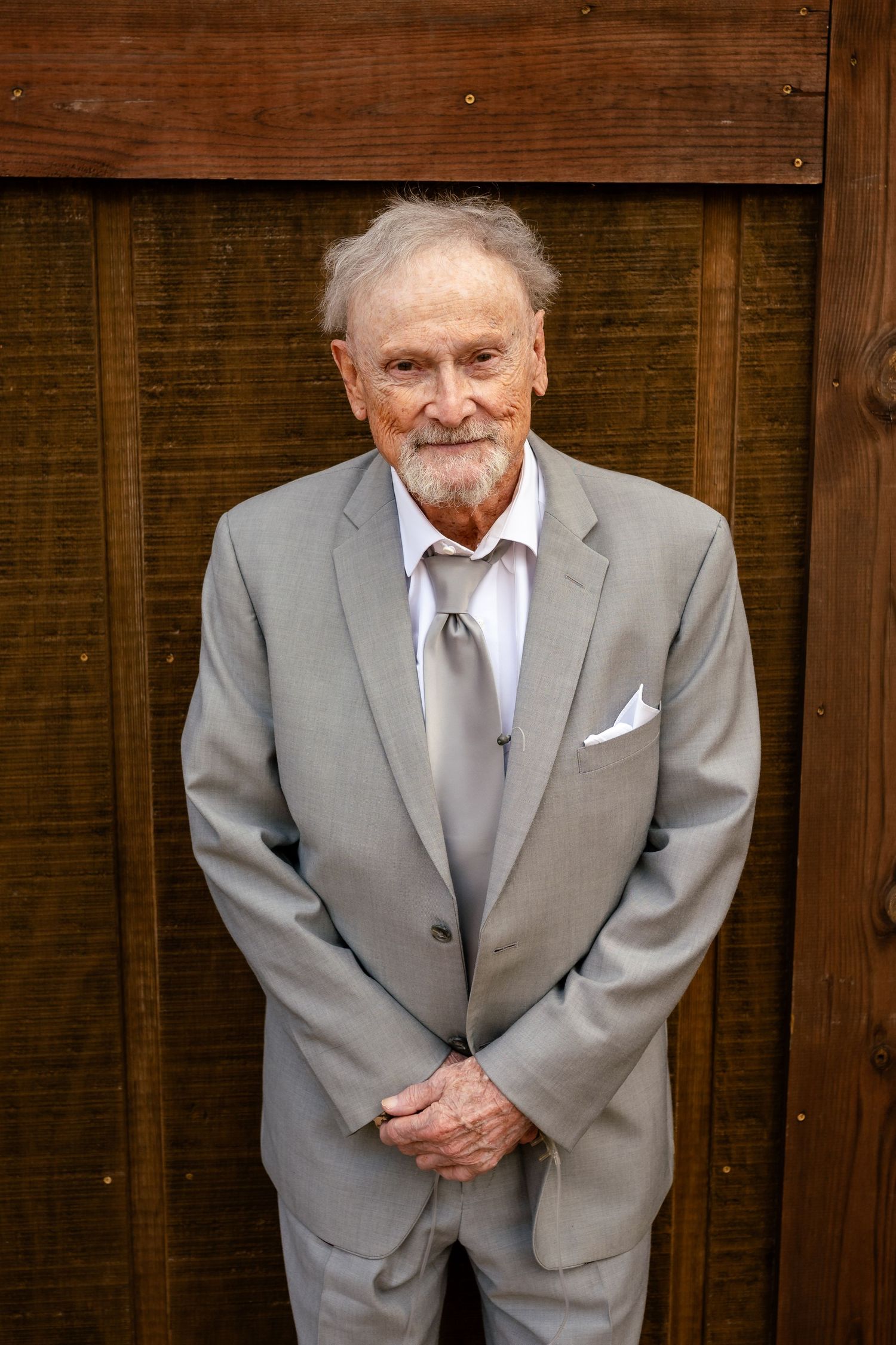 Elderly man in gray suit with tie and pocket square smiling warmly, standing against a wooden backdrop.