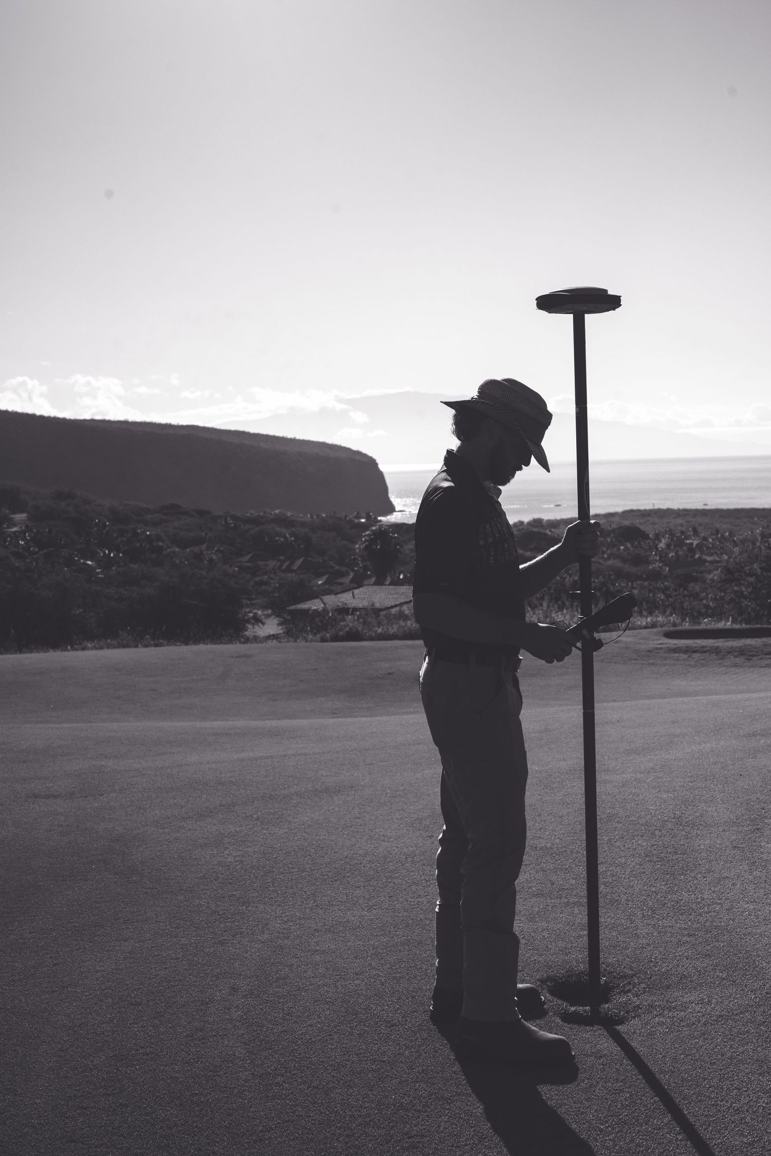 Silhouette of a surveyor holding a GPS pole on a road with scenic coastal hills in black and white.