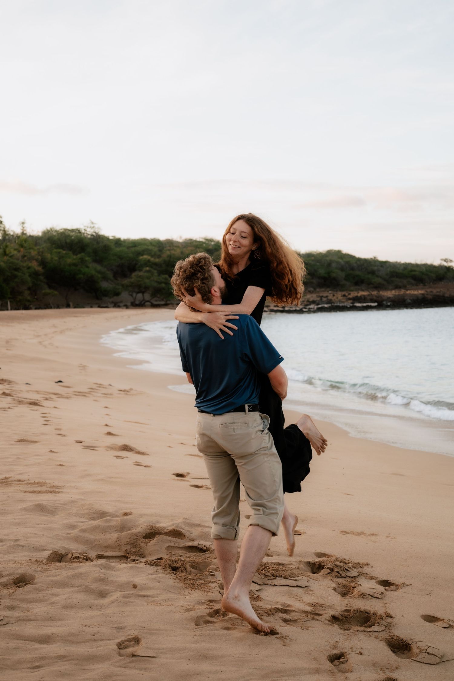 A couple embraces playfully on a sandy beach at sunset with ocean waves in the background.