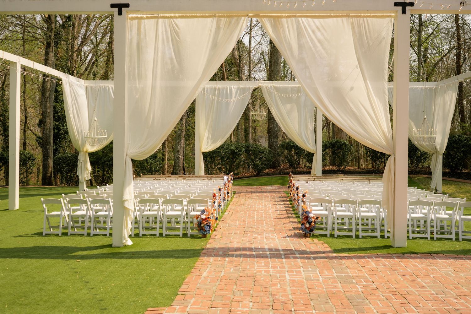 Elegant outdoor wedding ceremony setup with white draping canopy, rows of white chairs, and brick aisle on lush green lawn.