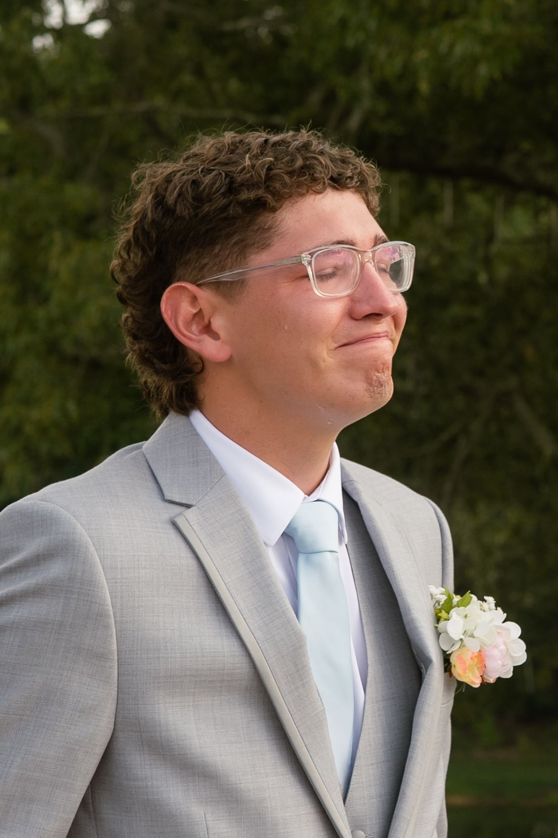 Young man in gray suit with light blue tie and boutonniere, wearing clear glasses, emotional expression outdoors.