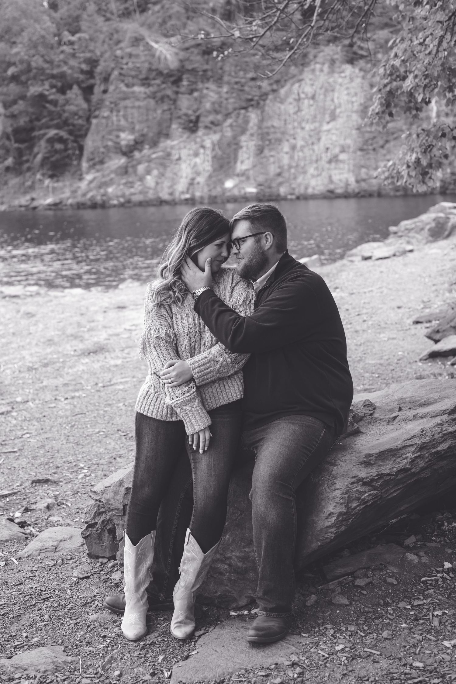 Black and white photo of a romantic couple sitting on rocks near a river, sharing an intimate moment outdoors.