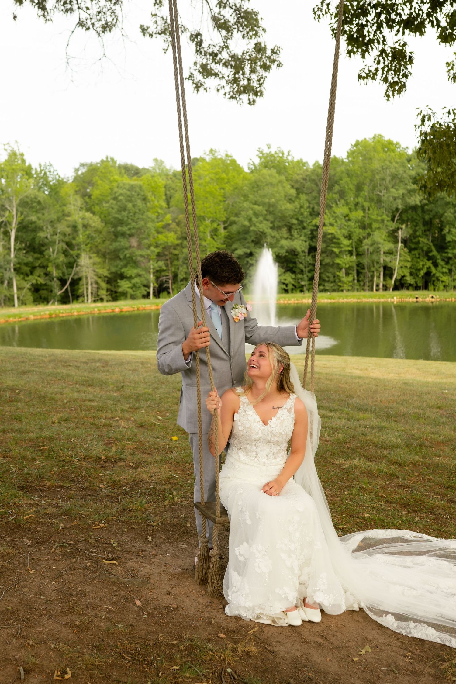 Groom in gray suit stands behind bride seated on tree swing near a serene pond with fountain backdrop.