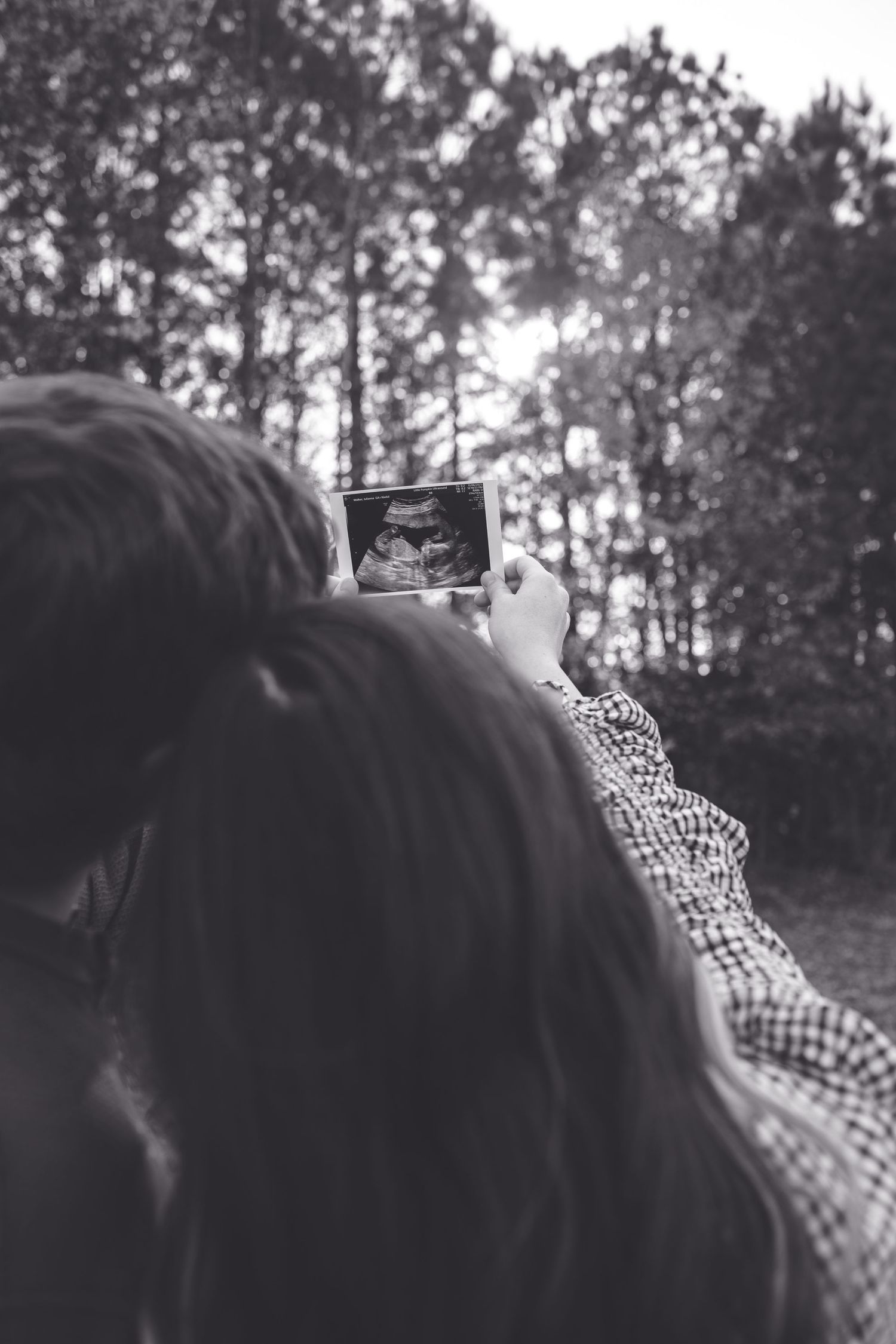 Couple holding ultrasound photo outdoors in black and white, surrounded by trees, announcing pregnancy.