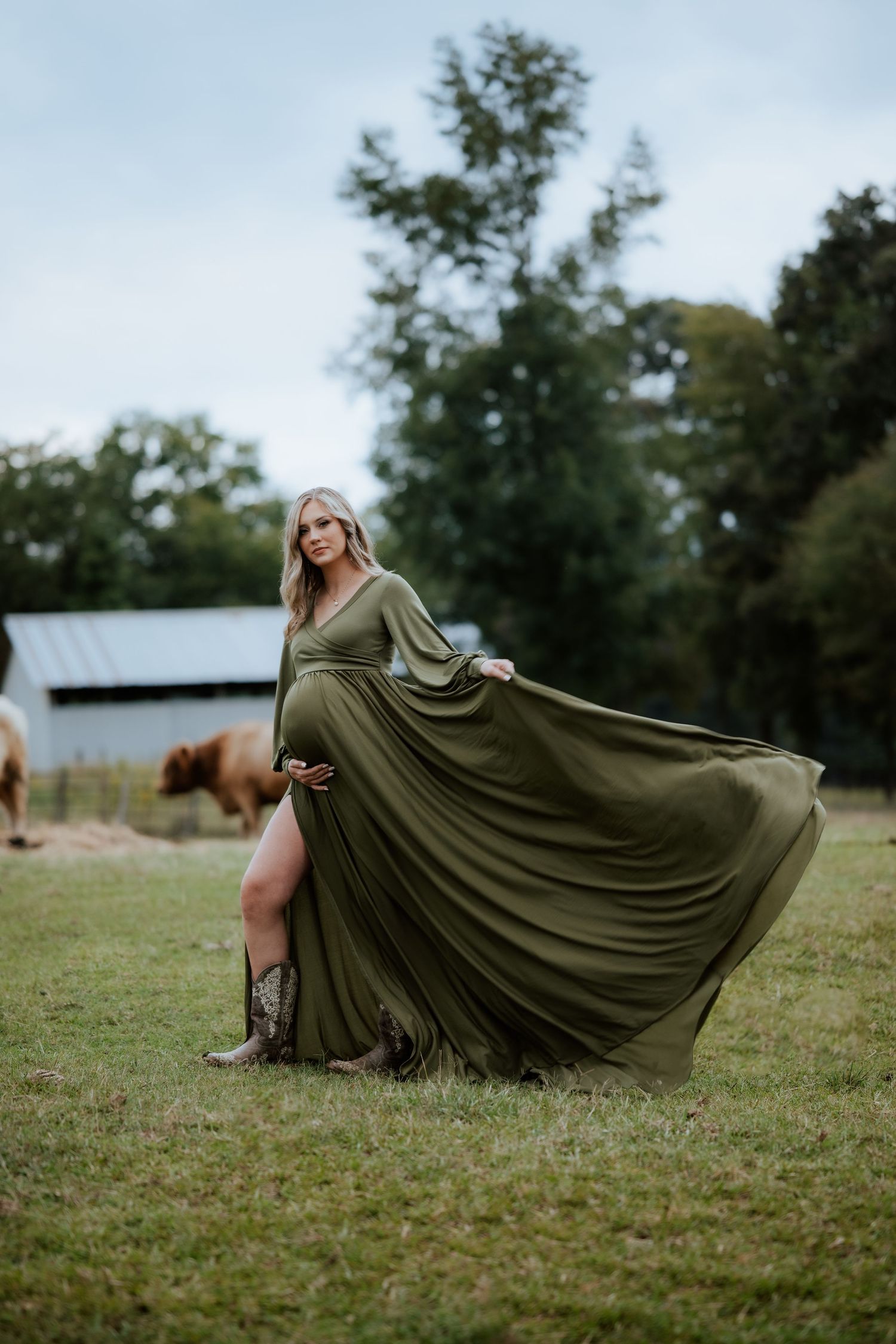 Dramatic maternity photo with flowing olive dress in rustic farm setting.