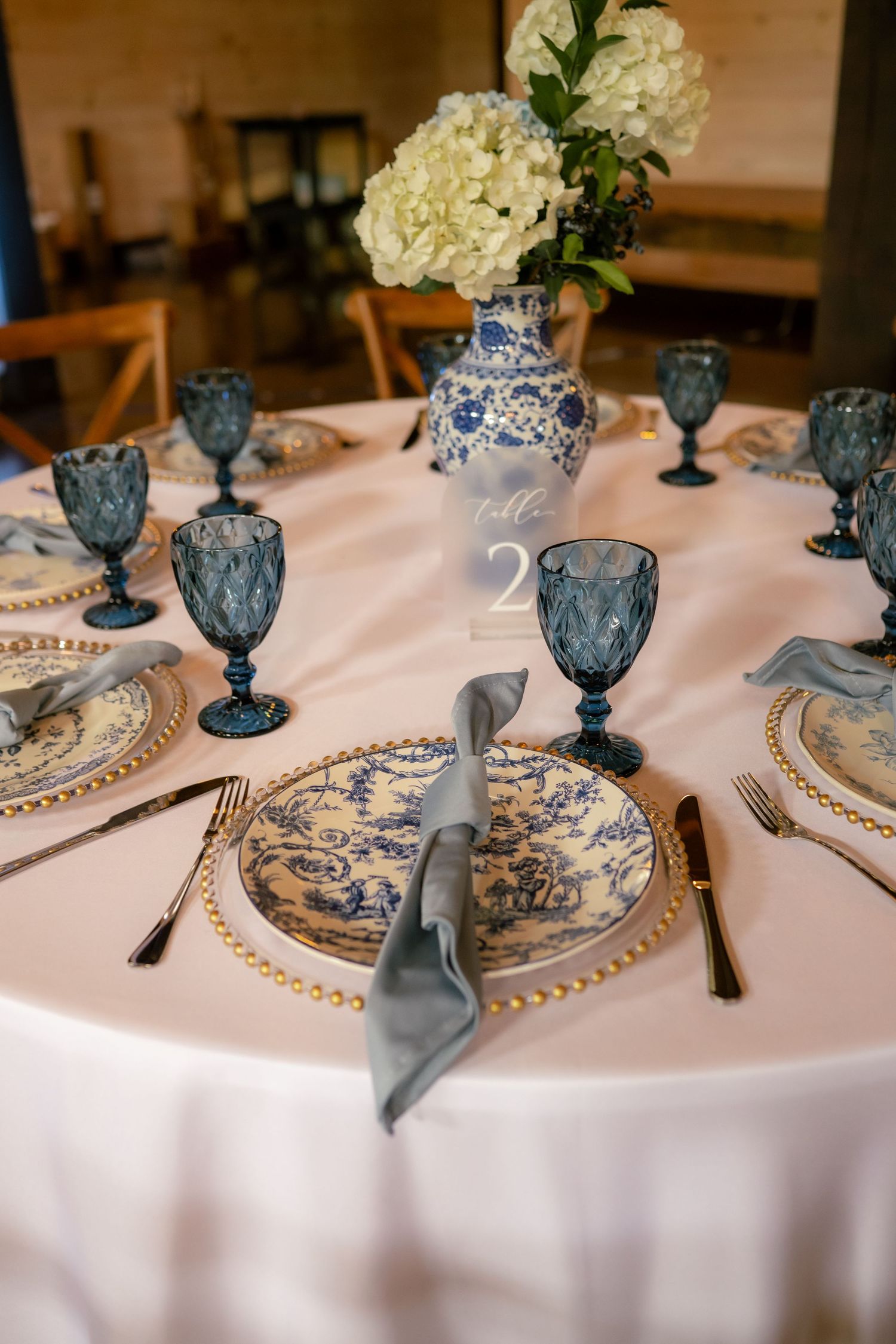 Elegant wedding table setting with blue toile plates, gold chargers, blue goblets, and white hydrangea centerpiece in blue vase.