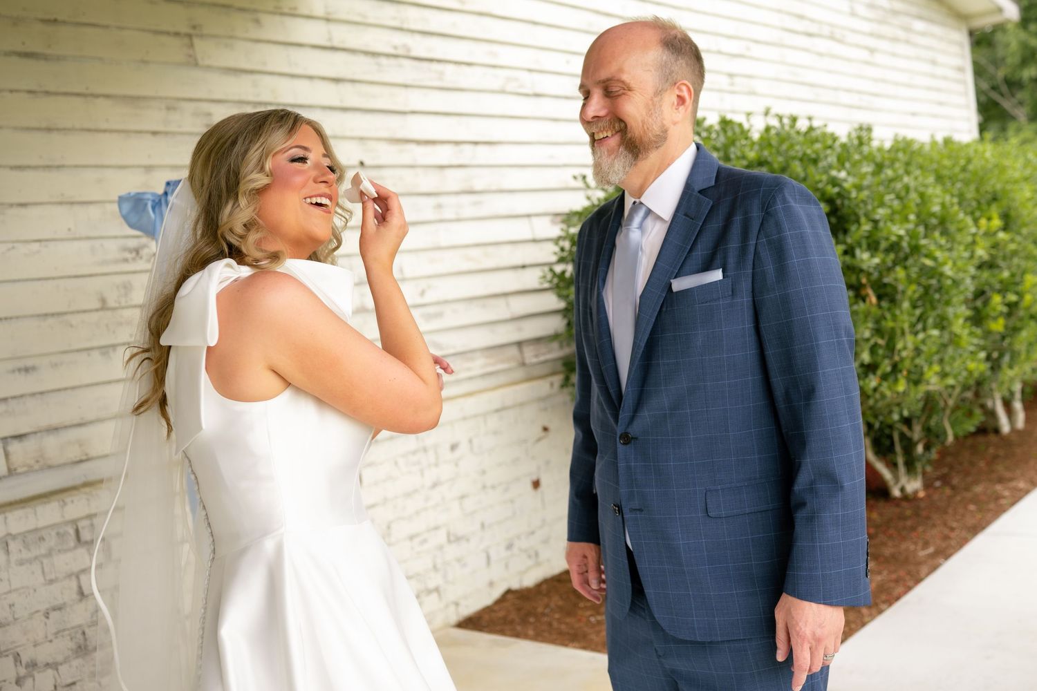 Bride in white dress laughing with groom in navy suit outdoors at wedding ceremony near white brick wall.