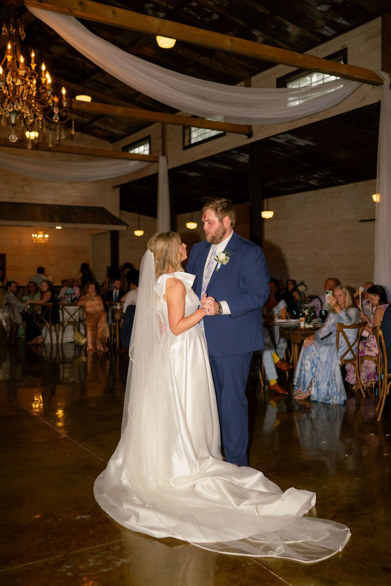 A bride and groom share their first dance in an elegant rustic barn reception hall adorned with chandeliers and draping.
