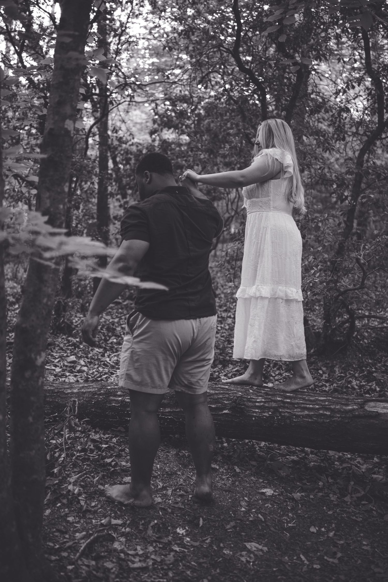 Black and white photo of a couple in the woods, woman in white dress balancing on a log while man steadies her.