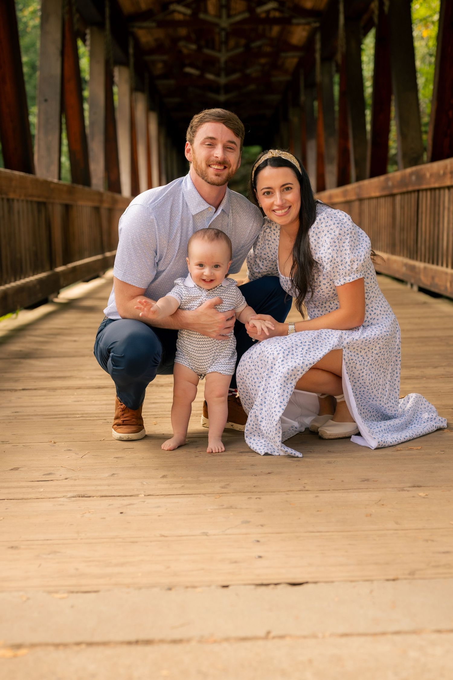 A family poses together on a wooden covered bridge walkway surrounded by green trees on a sunny day.