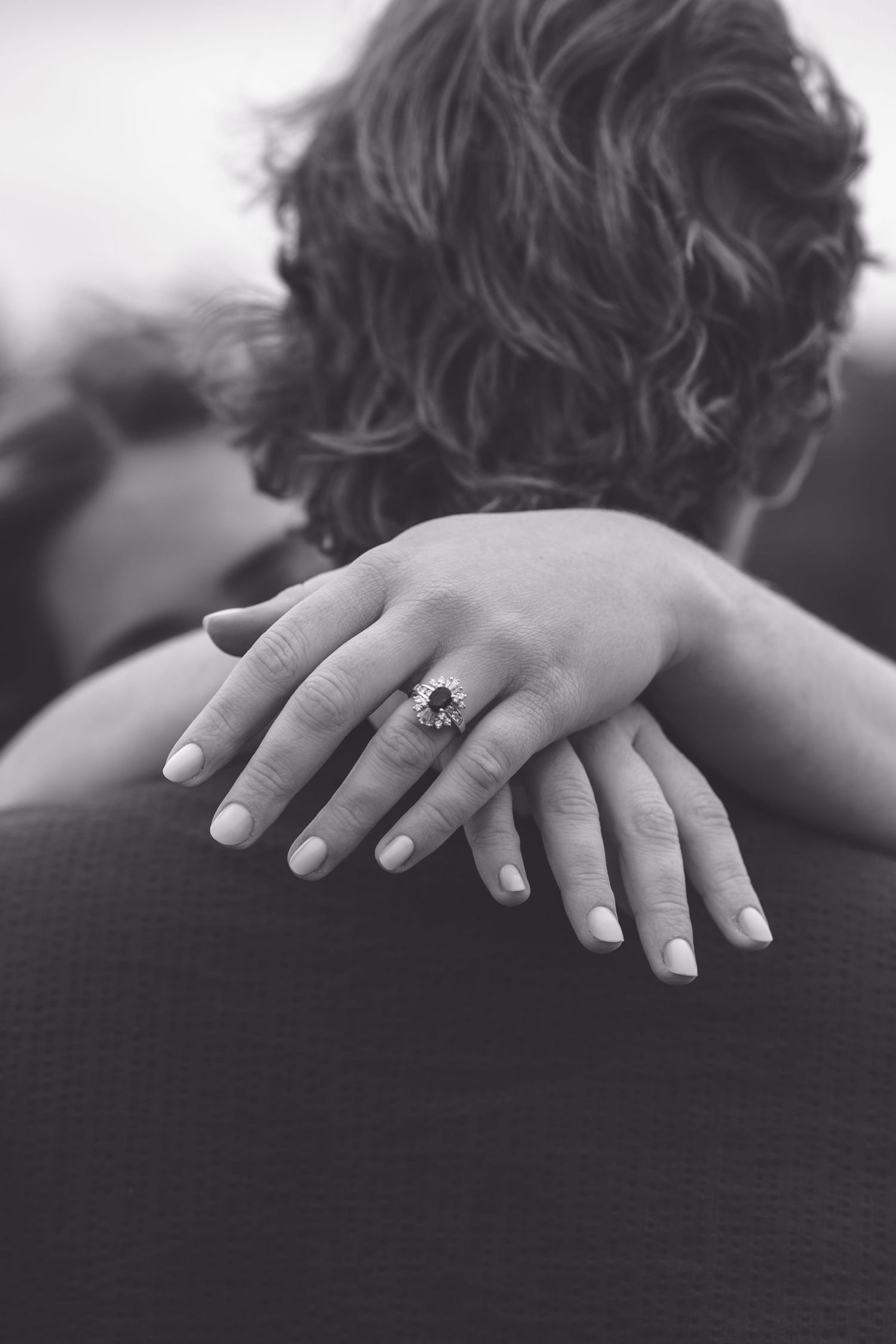 Black and white photo of a woman's hand with an engagement ring resting on a man's shoulder.