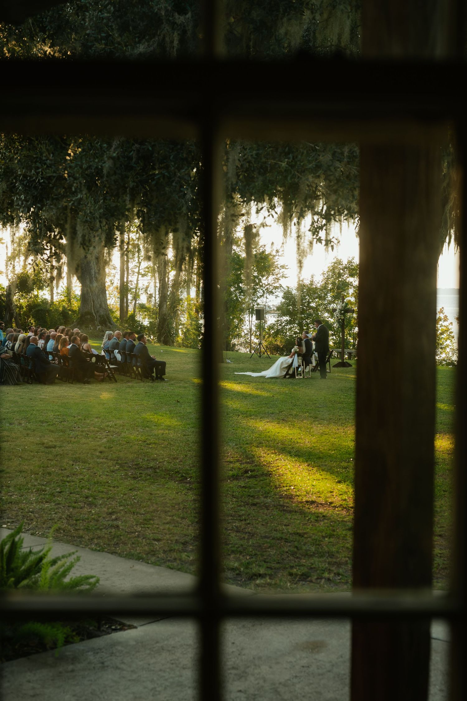 View through window of sunset wedding ceremony on grassy lawn with Spanish moss draped trees in background.