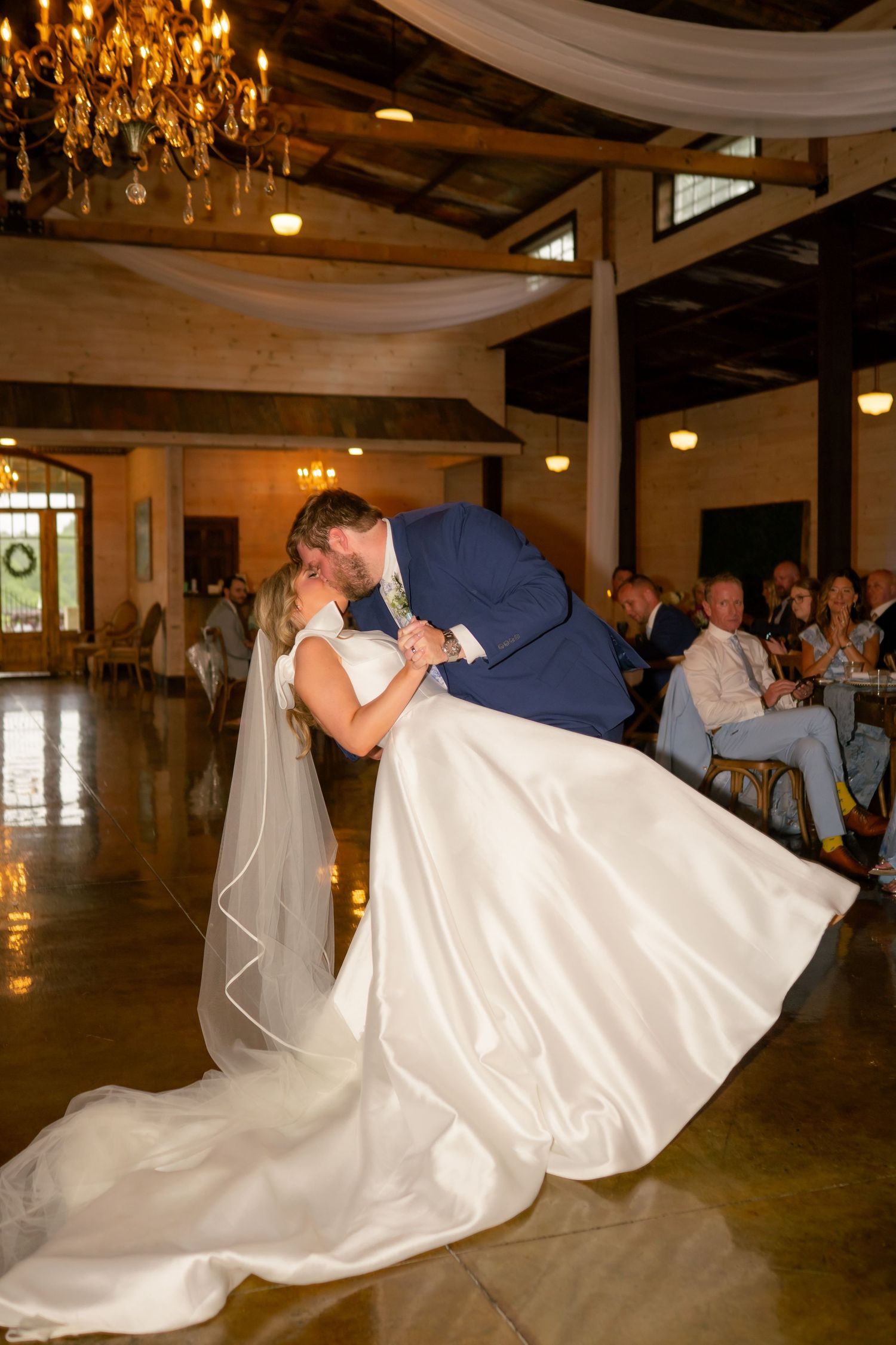 Bride and groom share a romantic dip kiss on the dance floor during their wedding reception at a rustic venue.