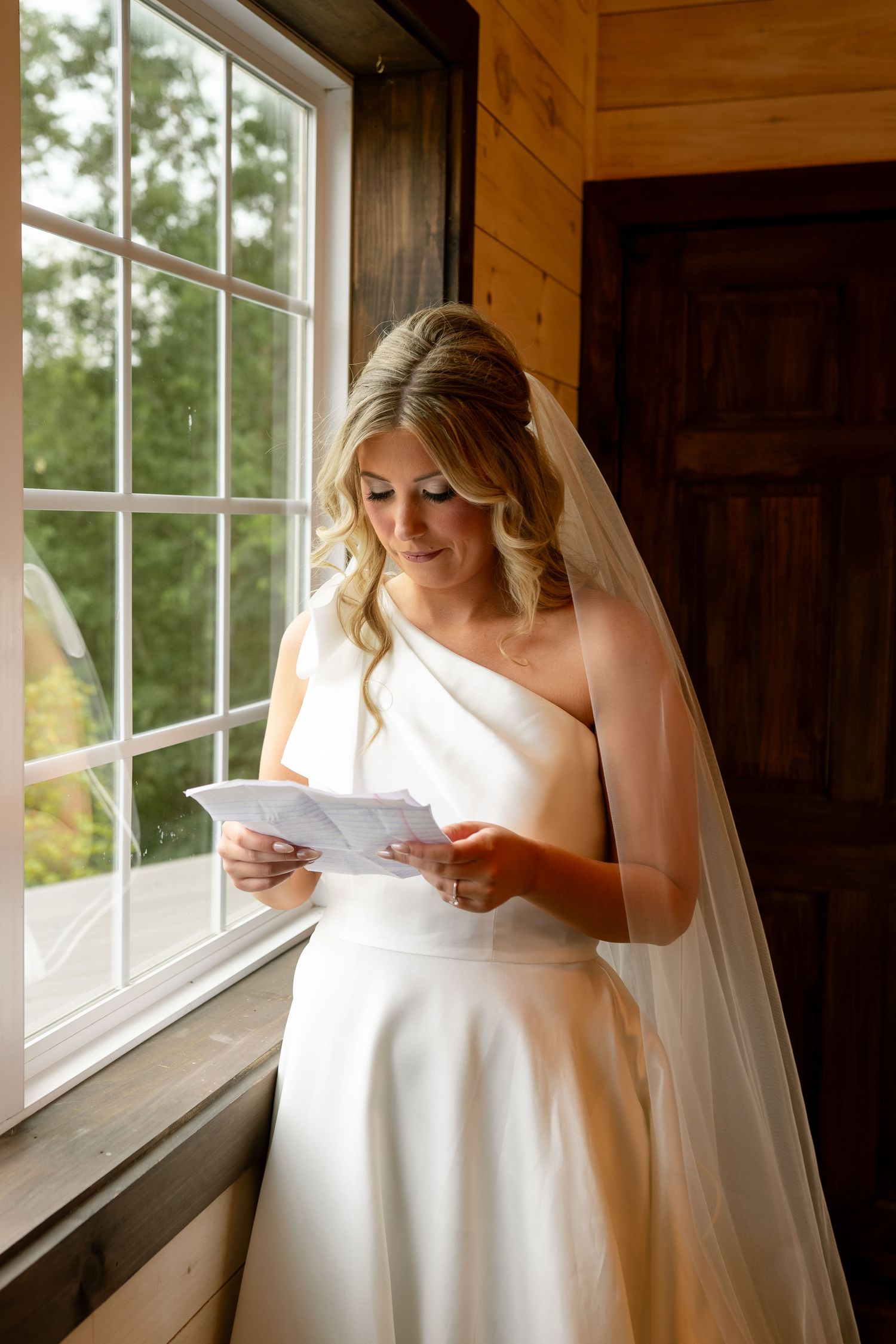 Bride in white one-shoulder gown and veil reads a letter by a sunlit window on her wedding day.